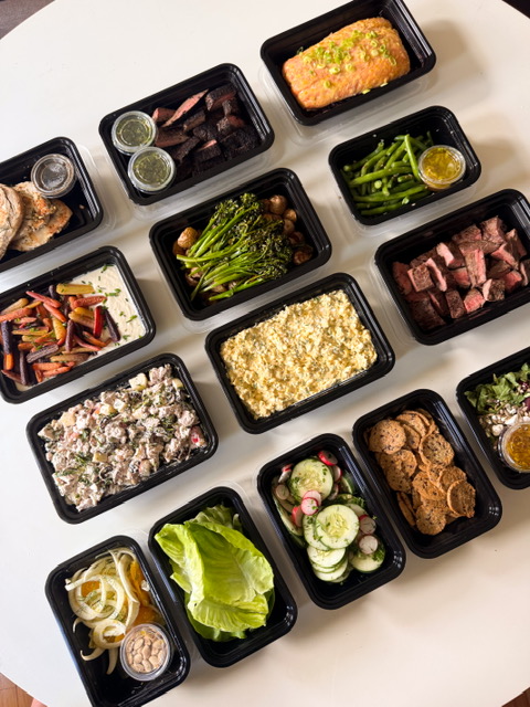 Multiple black food containers with various dishes on a white surface, including salads, cooked meats, and side dishes.