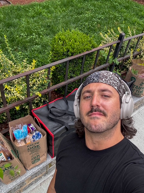 Man with headphones and bandana taking a selfie outdoors near a fence and green plants.