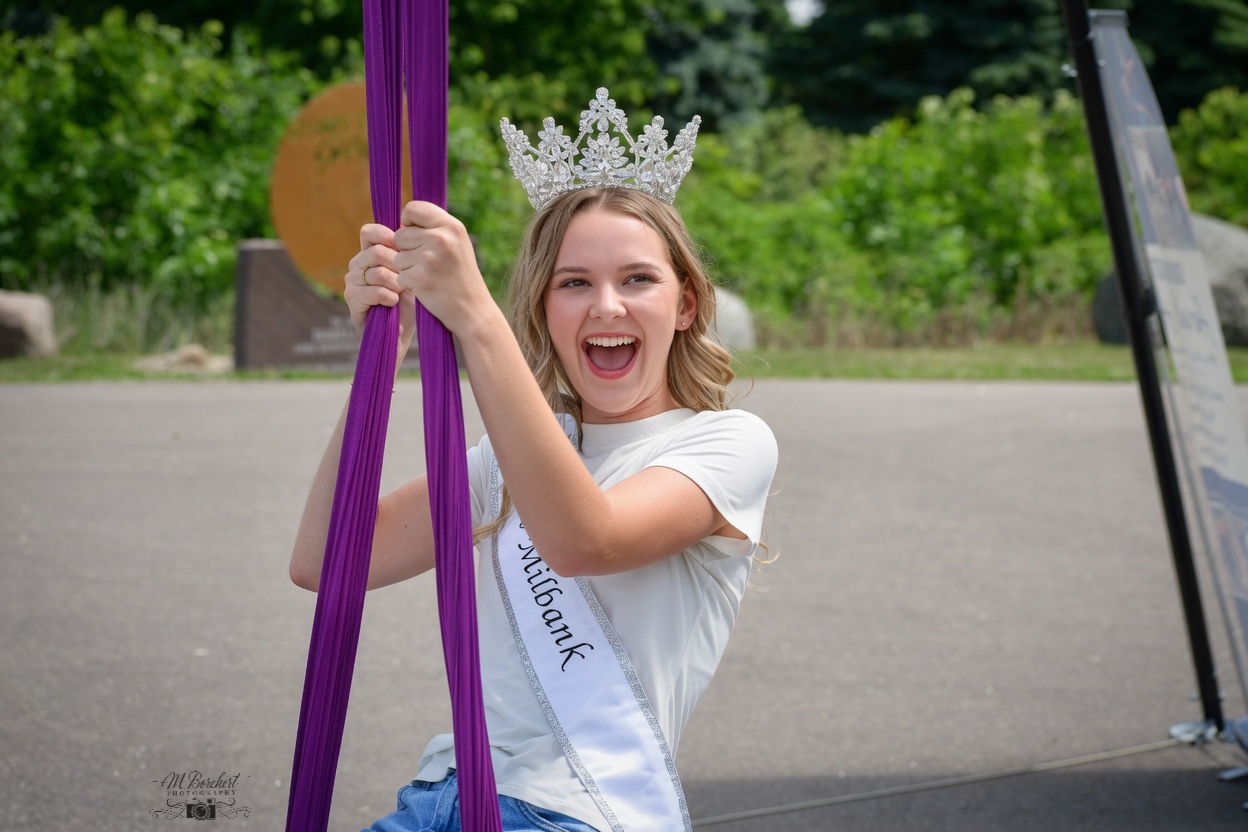 Young girl wearing a crown and sash, smiling and holding purple ribbons outdoors.