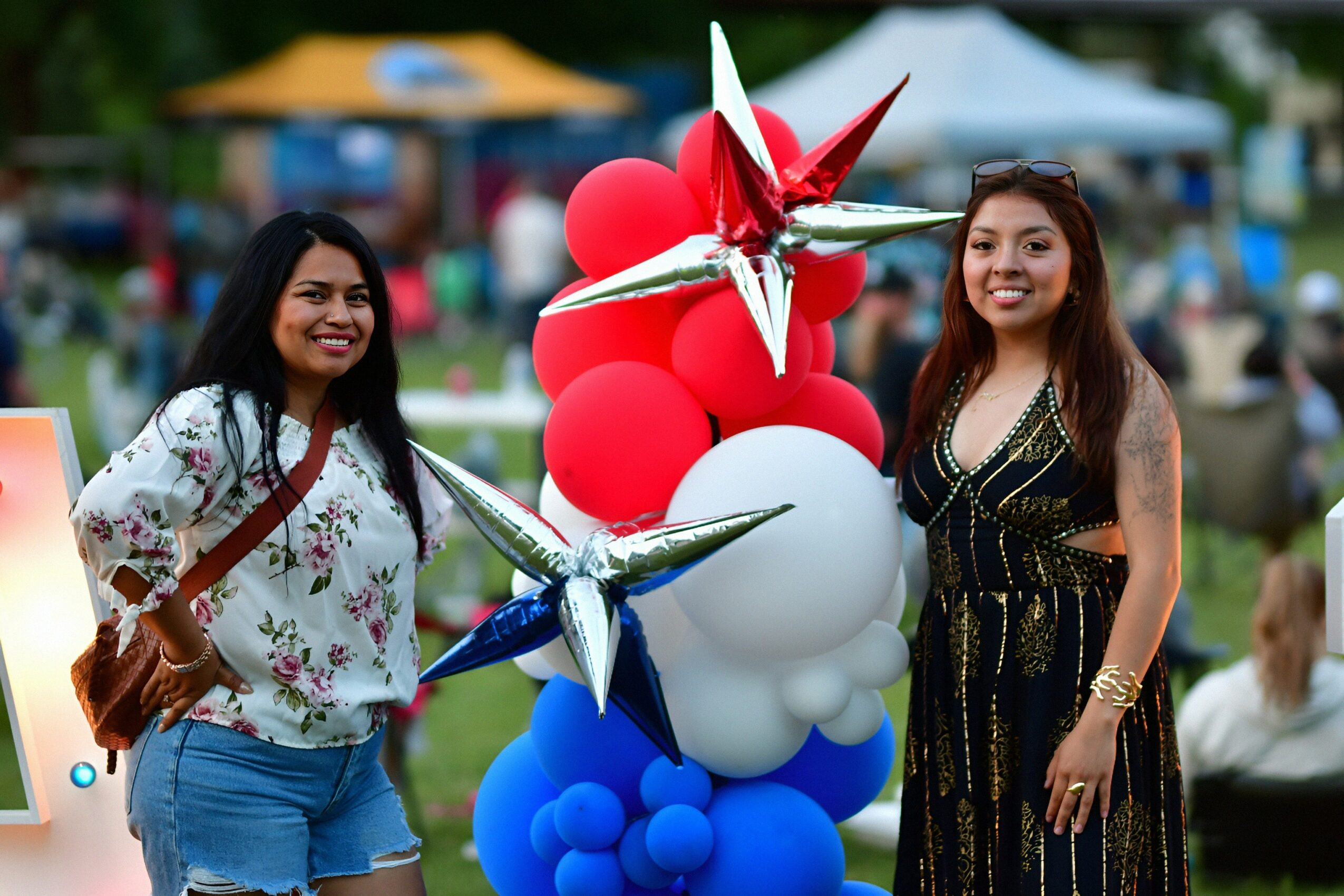 Two women stand outdoors near a colorful balloon arrangement with red, white, and blue balloons, smiling at the camera.