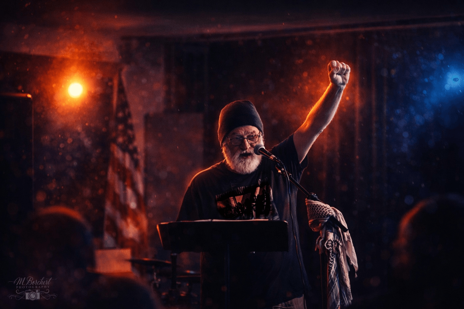 Person with a beard and glasses raising a fist on stage, with American flag in background, stage lights shining.