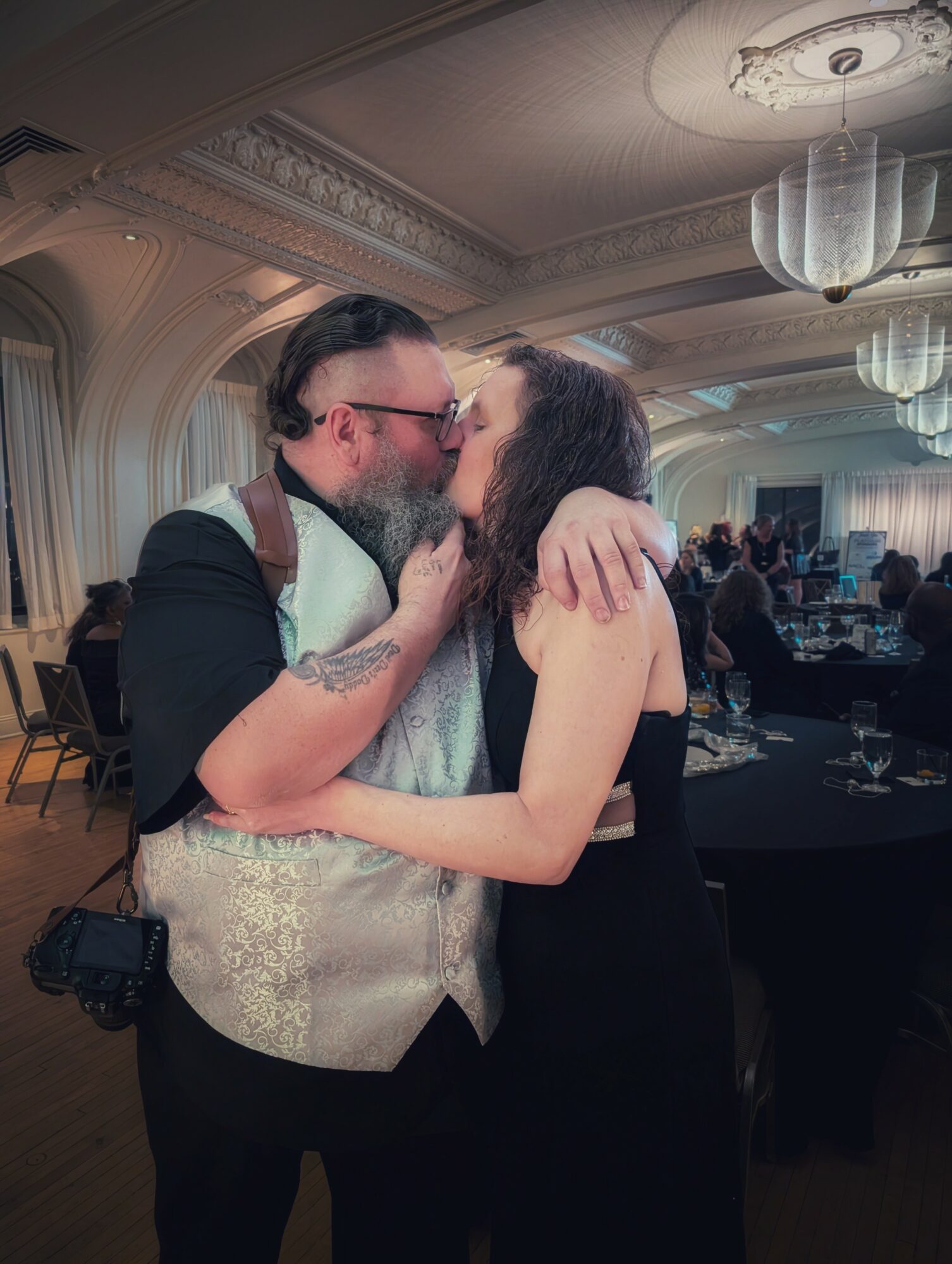 Two people kissing in a decorated ballroom with chandeliers and seated guests, one with glasses and a beard, the other with long hair.