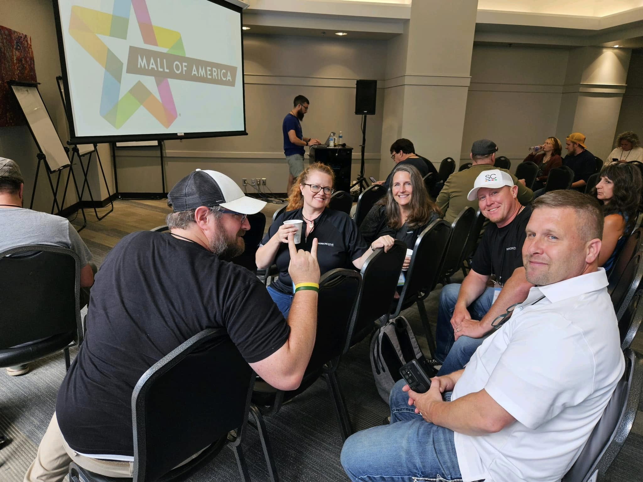 Group of people seated in a conference room, some using phones, with a large screen displaying 'Mall of America' in the background.