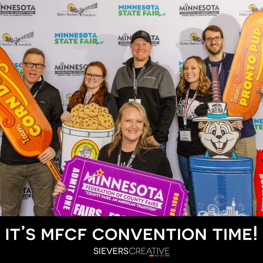 Group of six people smiling at Minnesota State Fair event, holding colorful signs and props, with a backdrop of fair logos.