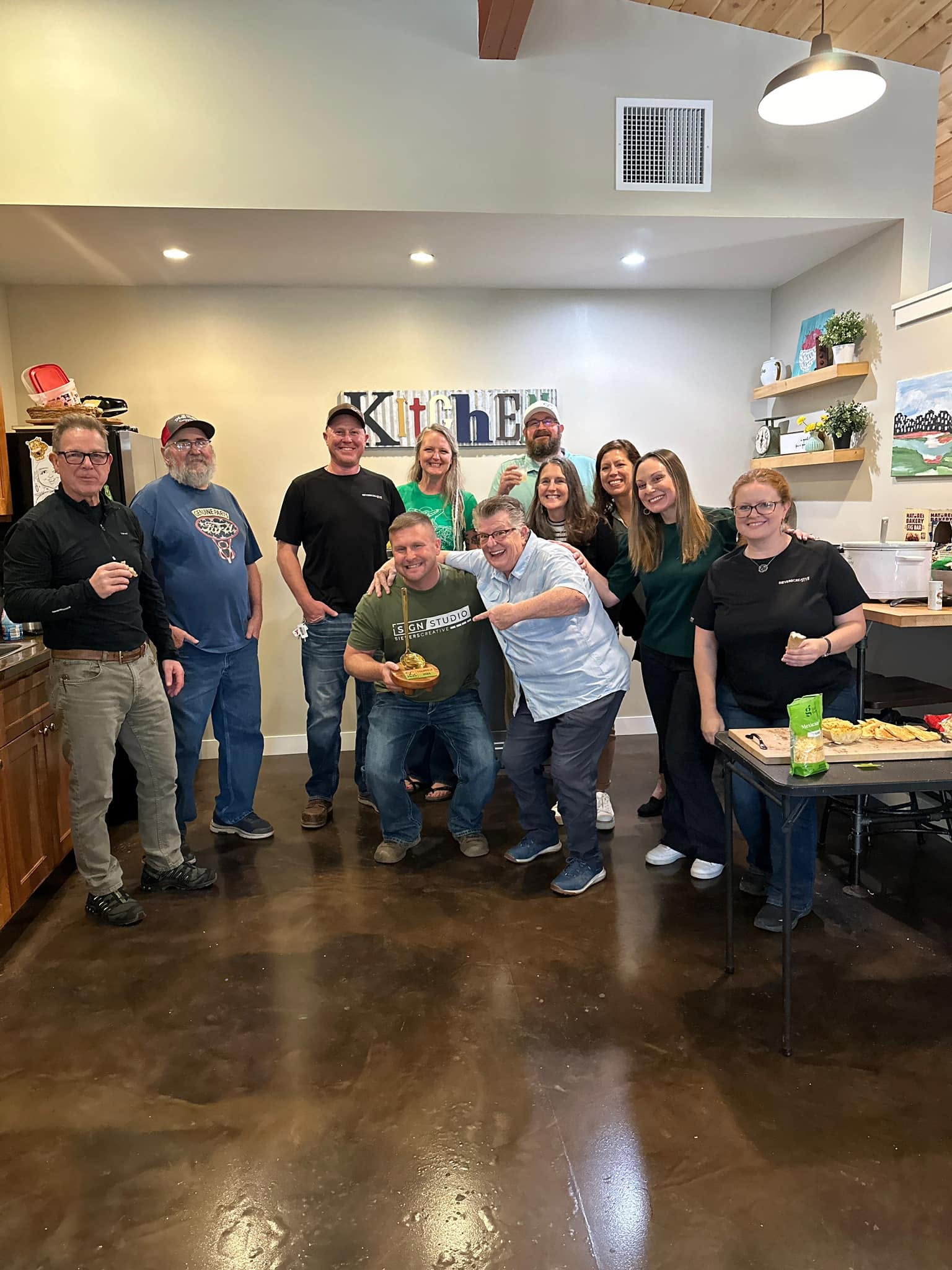 Group of ten people standing indoors, some holding food, smiling, in a room with shelves and decorations.