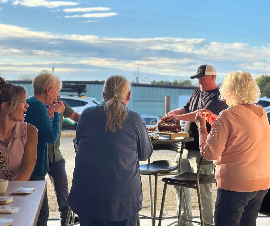 Group of five people standing outdoors near a body of water, with a man serving food, under a partly cloudy sky.