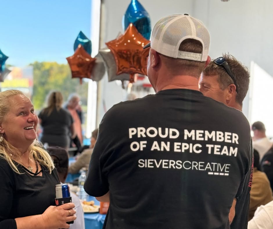 Person wearing a black T-shirt with text, talking to smiling woman holding a drink, decorated with balloons in background.