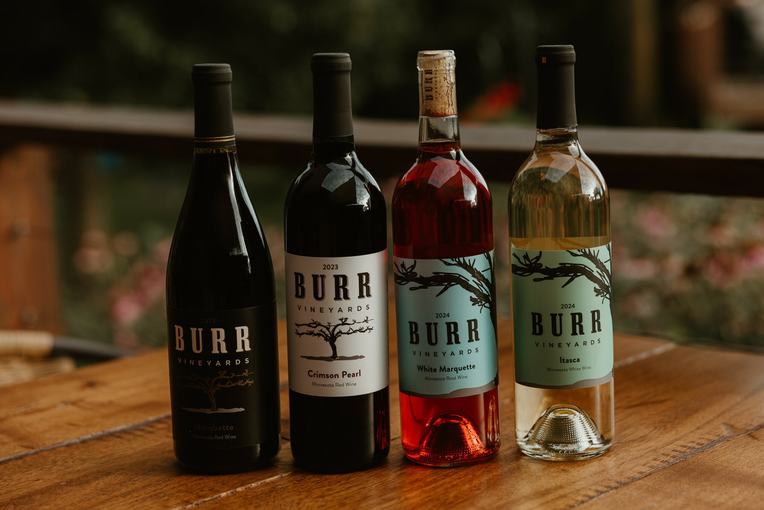 Four bottles of wine or spirits on a wooden surface with a blurred outdoor background.