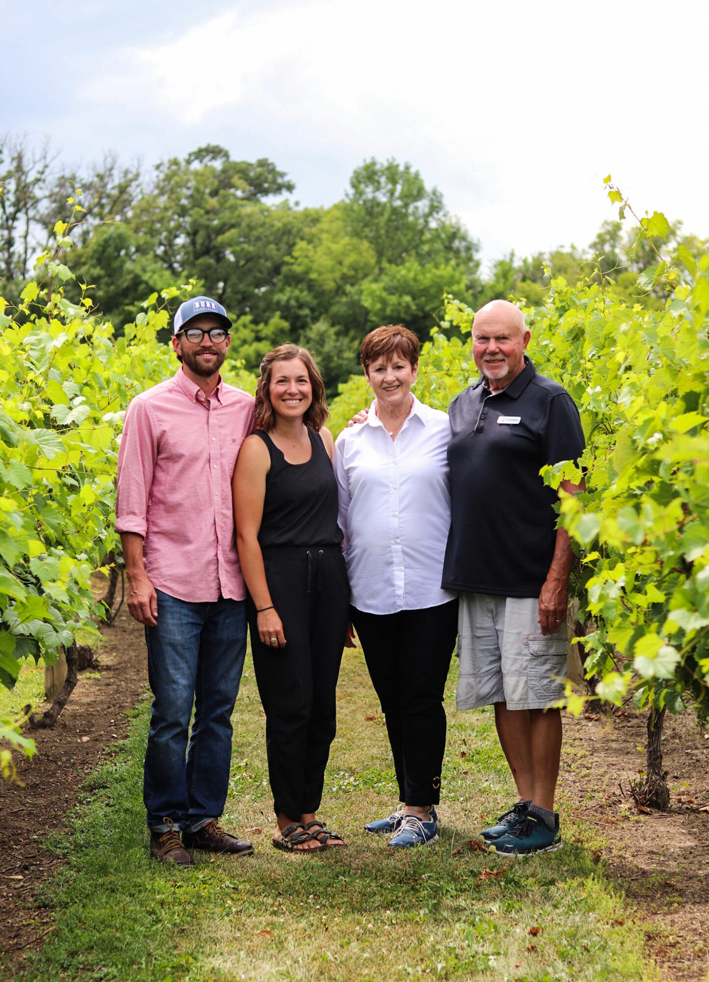 Four people standing in a vineyard with green vines and trees in the background, smiling at the camera.