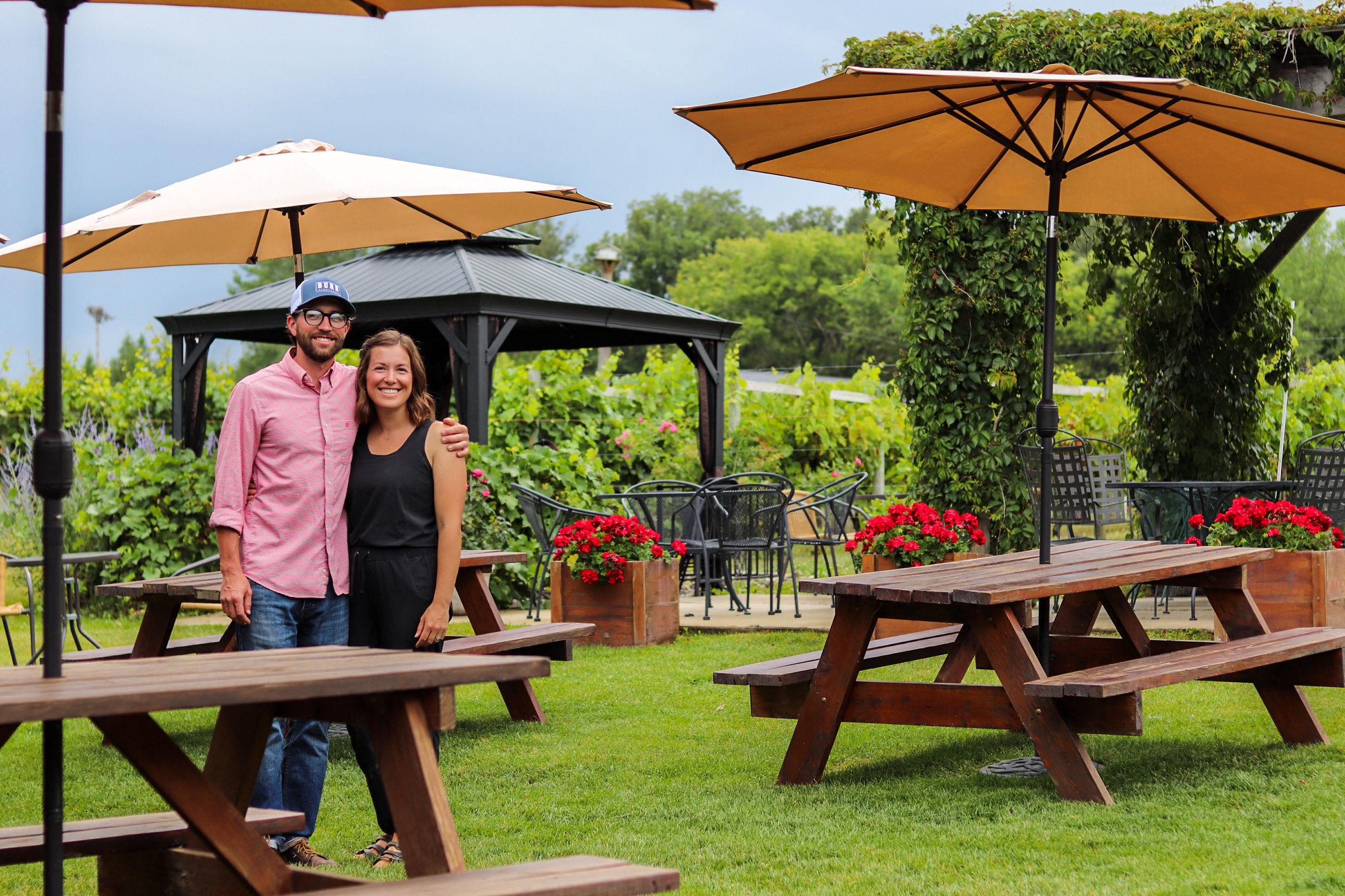 A man and woman stand together outdoors near picnic tables and umbrellas, with greenery and a gazebo in the background.
