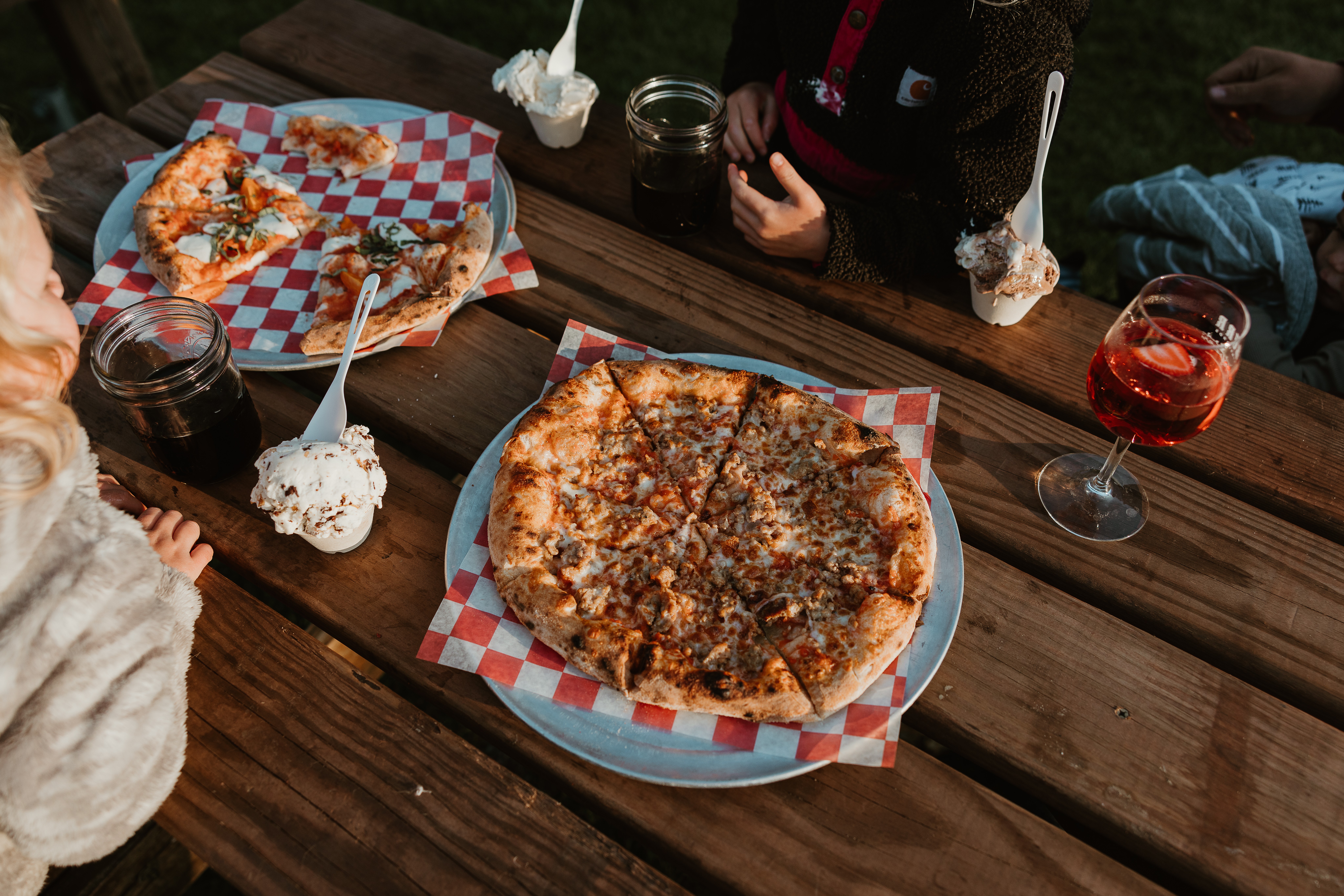 Pizza on a checkered paper tray, drinks, and desserts on a wooden table outdoors.