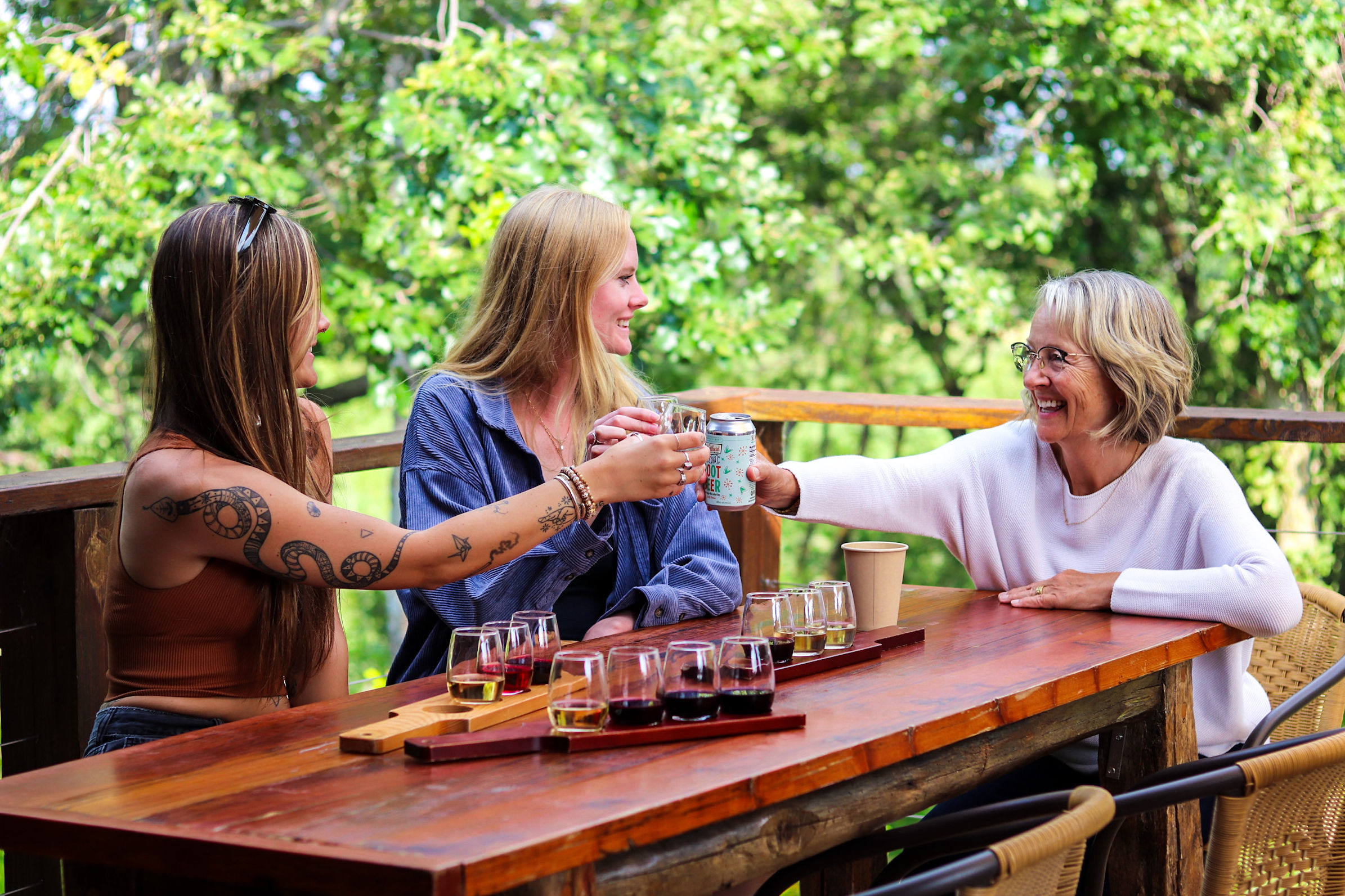 Three women sitting at a wooden table outdoors, smiling, with glasses and a tray of drinks, surrounded by green trees.