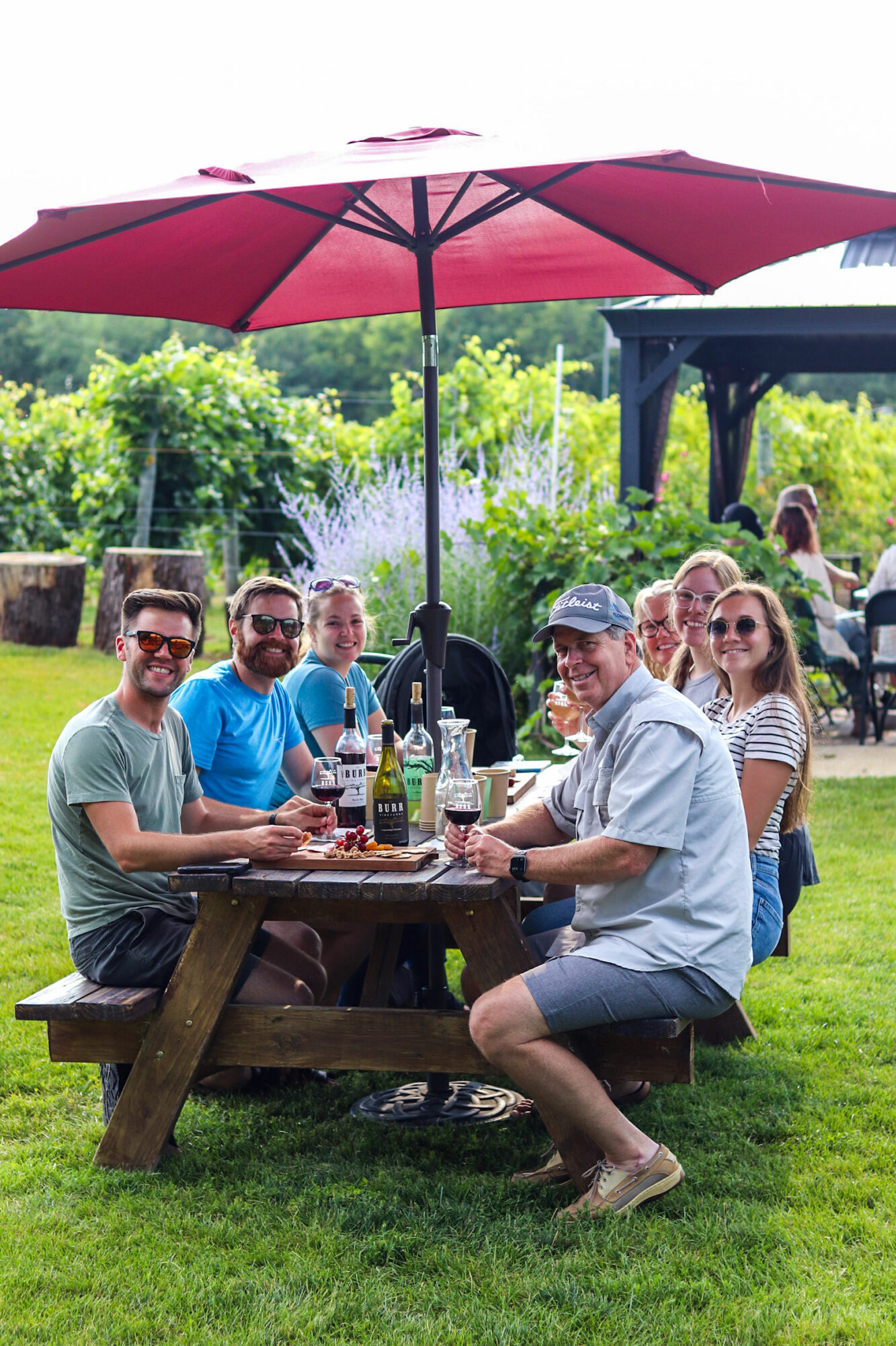 Group of six people sitting at a picnic table outdoors under a large pink umbrella, smiling and enjoying drinks.