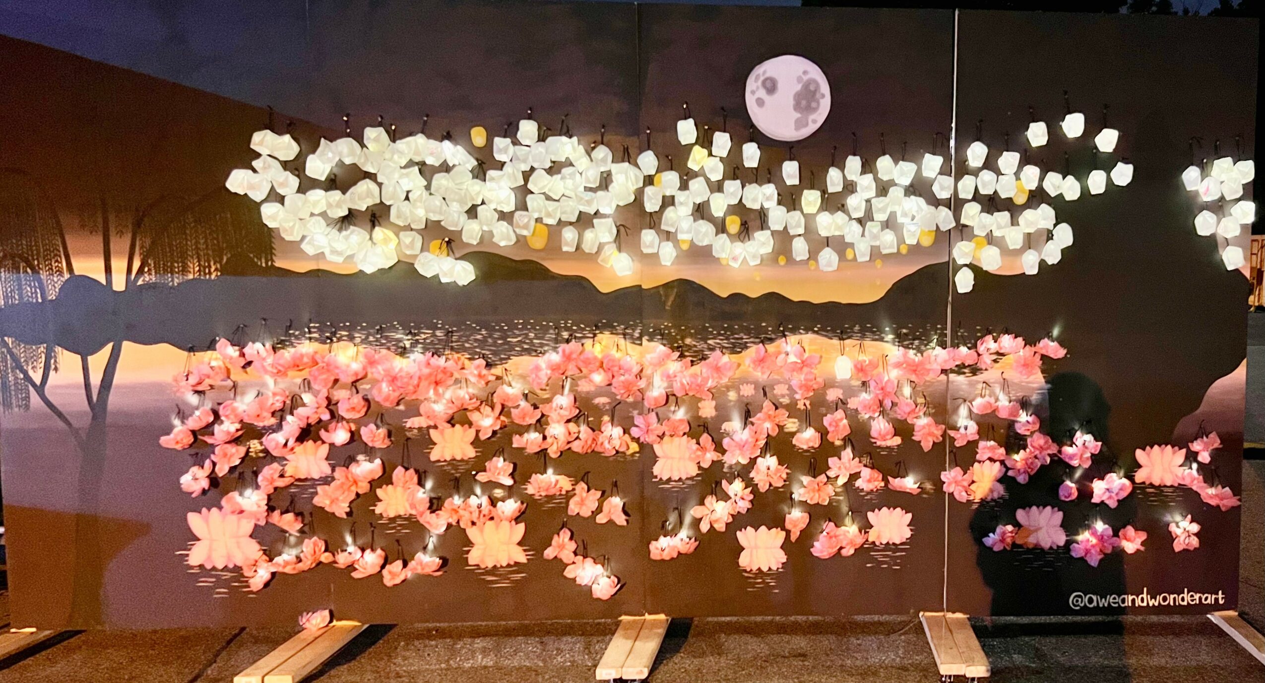 Display of illuminated clouds and a full moon, with pink and white flowers below, against a dark background.