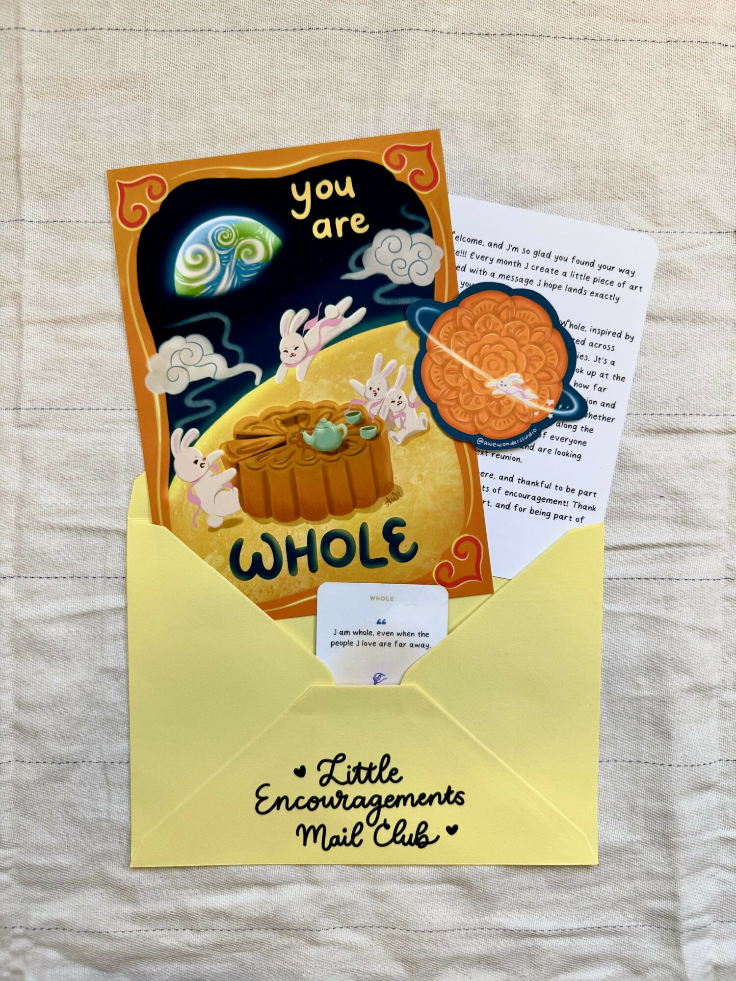Open yellow envelope with a card featuring a whimsical scene of rabbits, clouds, and a rainbow, and a round orange sticker.