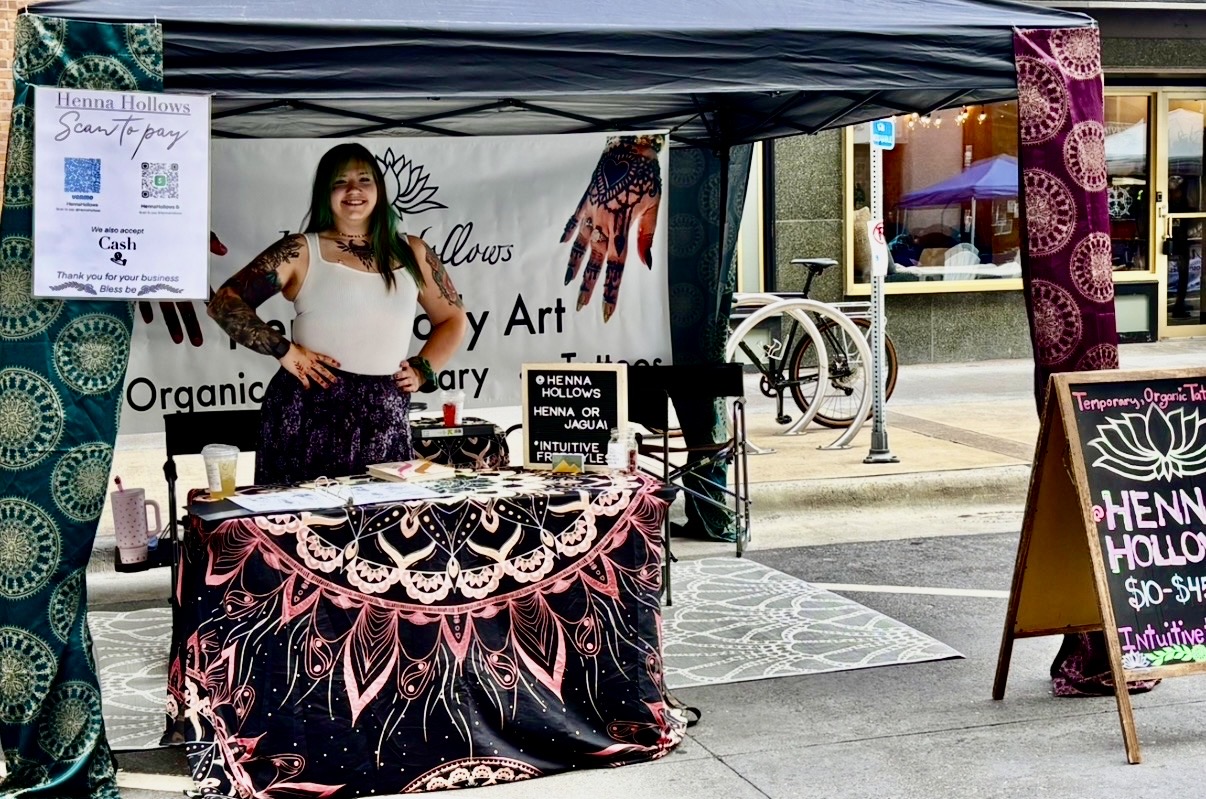 Woman standing behind a table with artwork and signs at an outdoor art booth, with a bicycle and storefront in background.