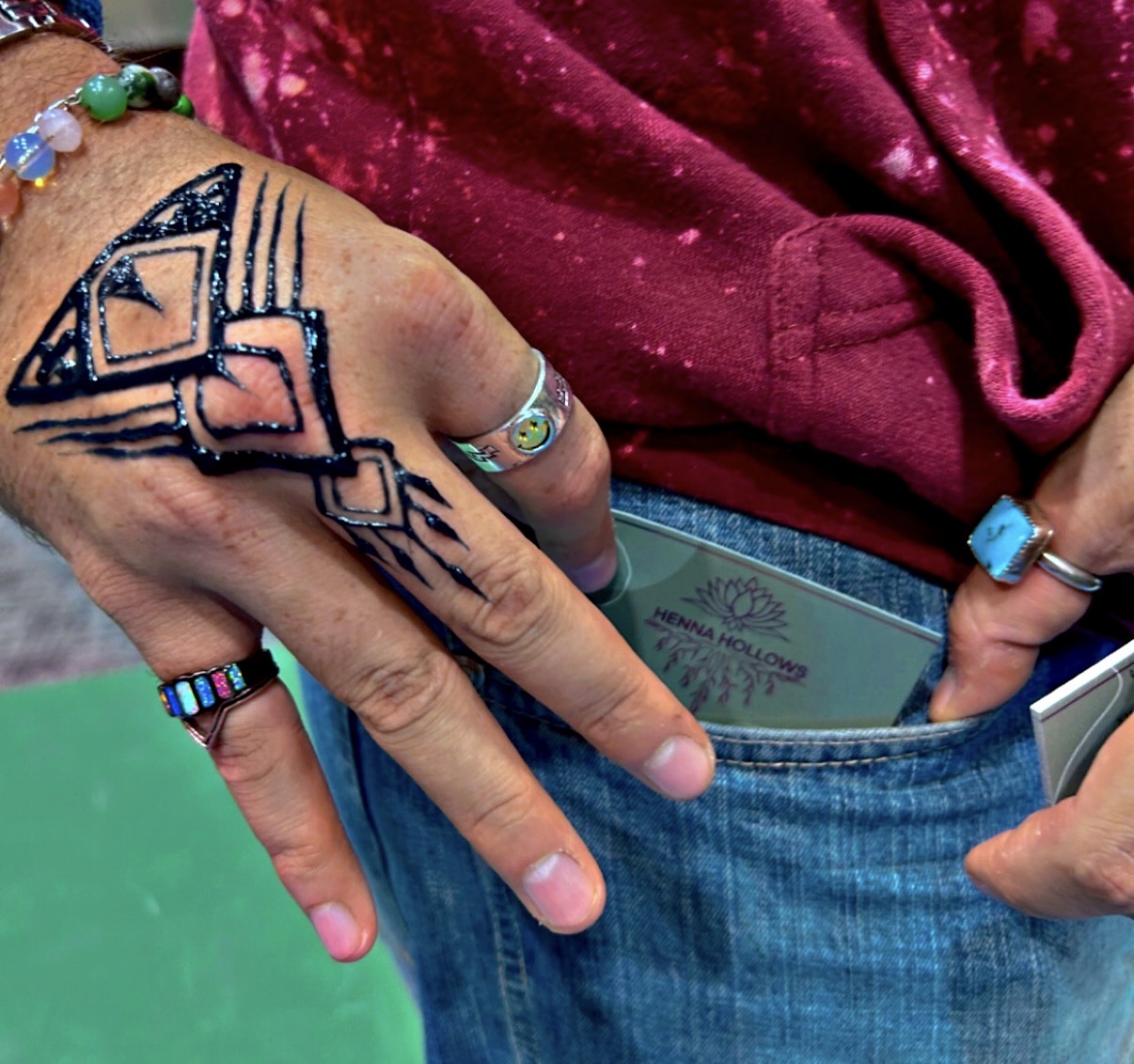 Person's hand with geometric tattoos, wearing colorful rings and bracelets, holding a phone and a wallet in front of a green background.
