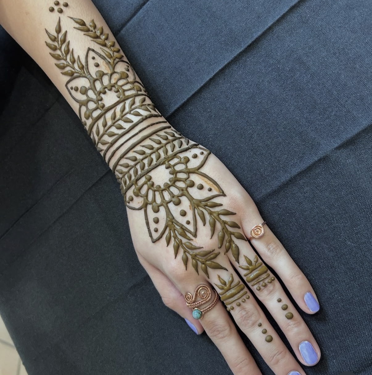Hand with henna design, rings on fingers, and painted nails, resting on a dark surface.