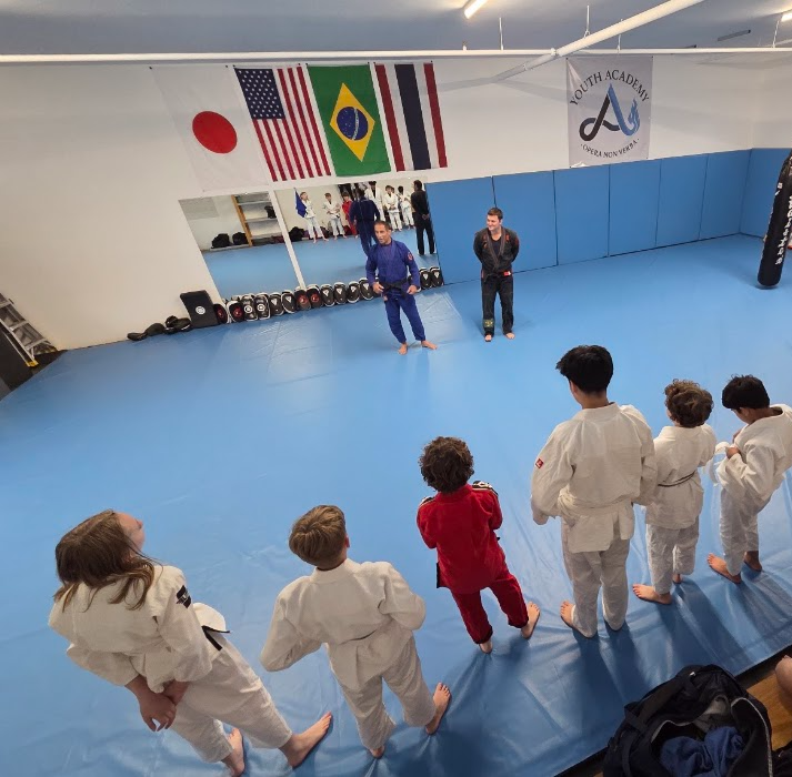 Children in martial arts uniforms standing on blue mat, facing instructors in a training room with flags and mirrors.
