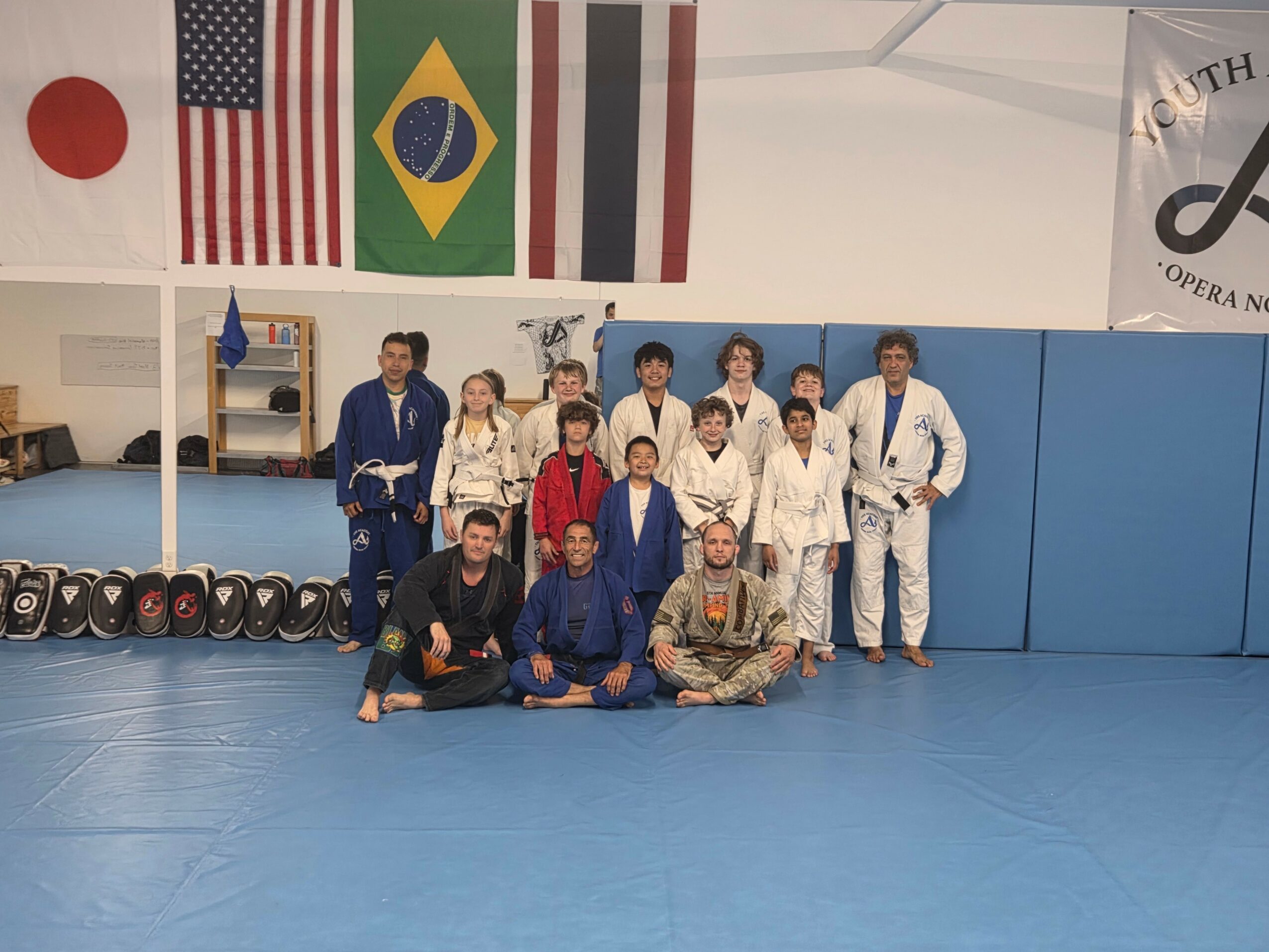 Group of people in martial arts uniforms and casual clothing posing in a gym with flags and mats.