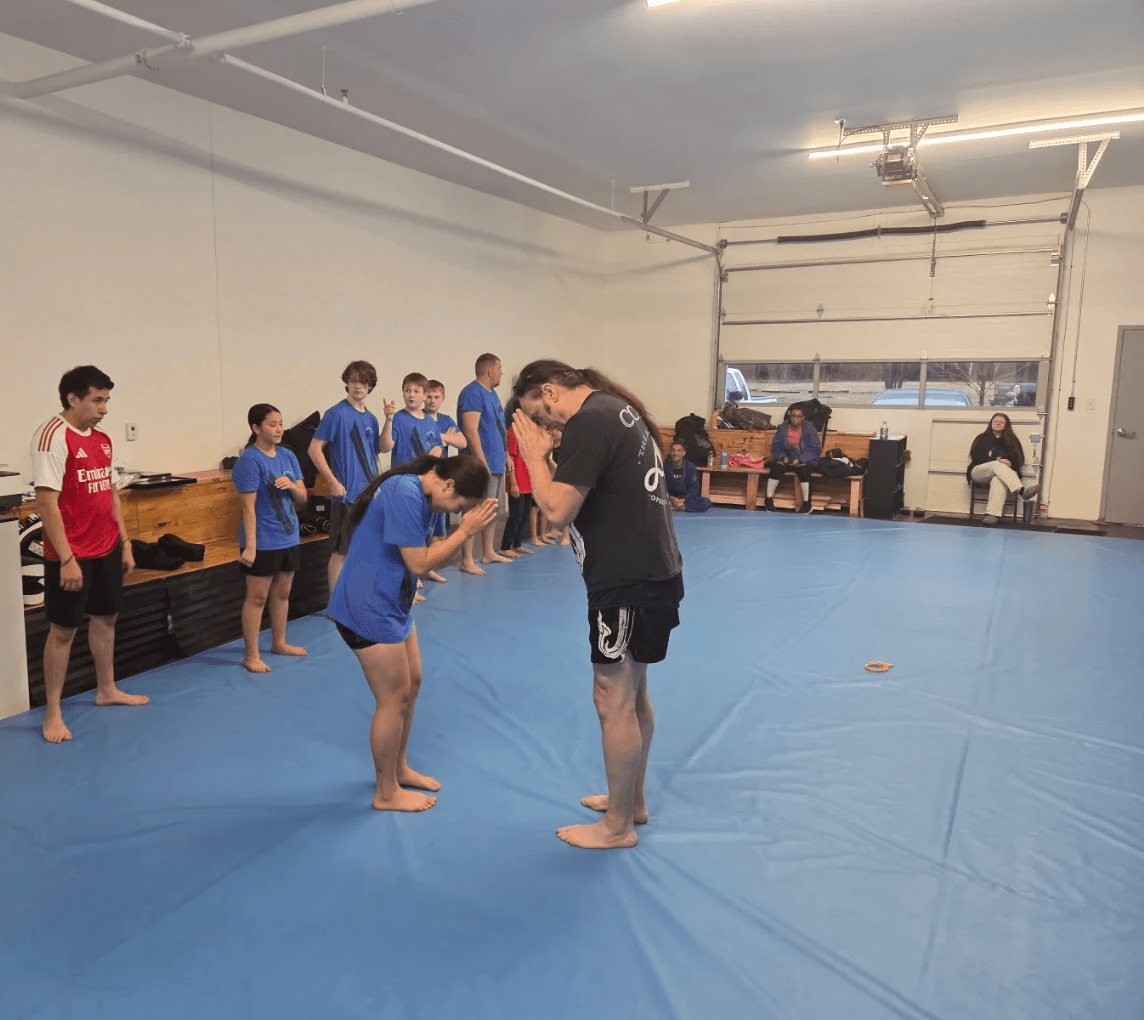 Instructor demonstrating a technique to children in a gym with blue mats, some sitting on benches, others standing, in a spacious room.