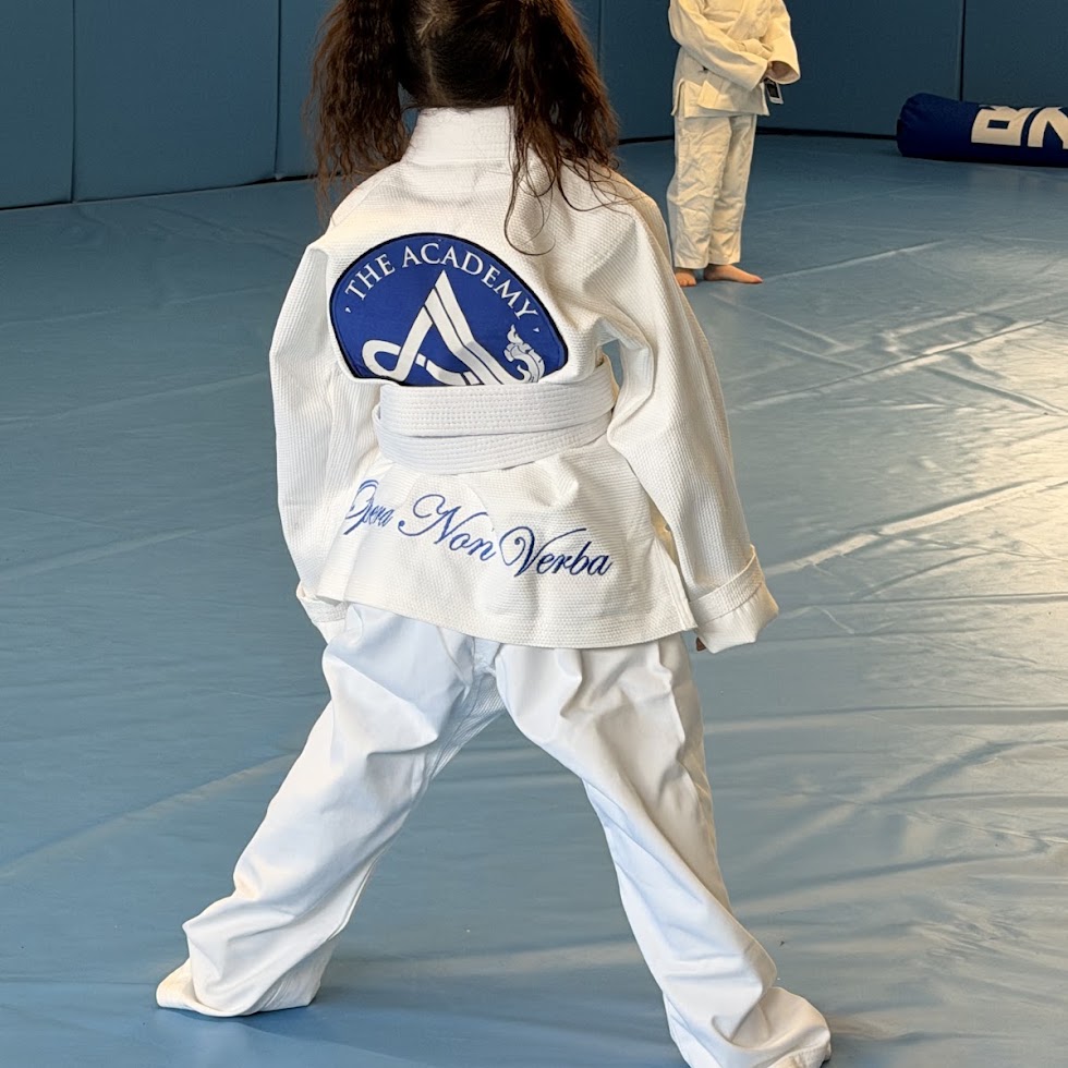 Child in a martial arts uniform standing on a mat, back view, with a logo on the back of the uniform.