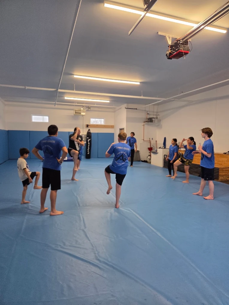 People in a martial arts training room, some standing and some practicing, with a blue padded floor and ceiling lights.
