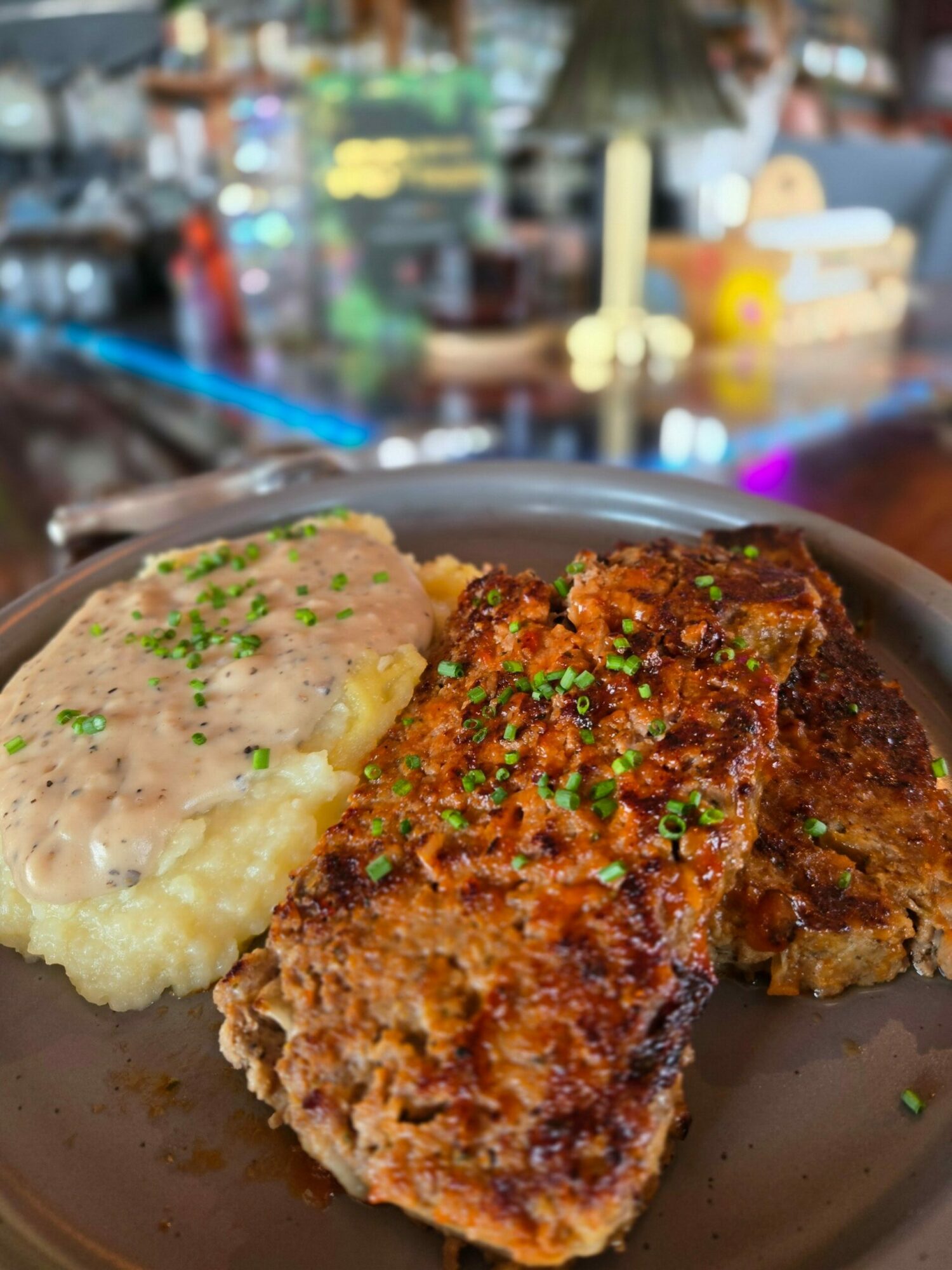 Piece of fried chicken with green herbs on mashed potatoes in a bowl, background of a busy restaurant. Word count: 20
