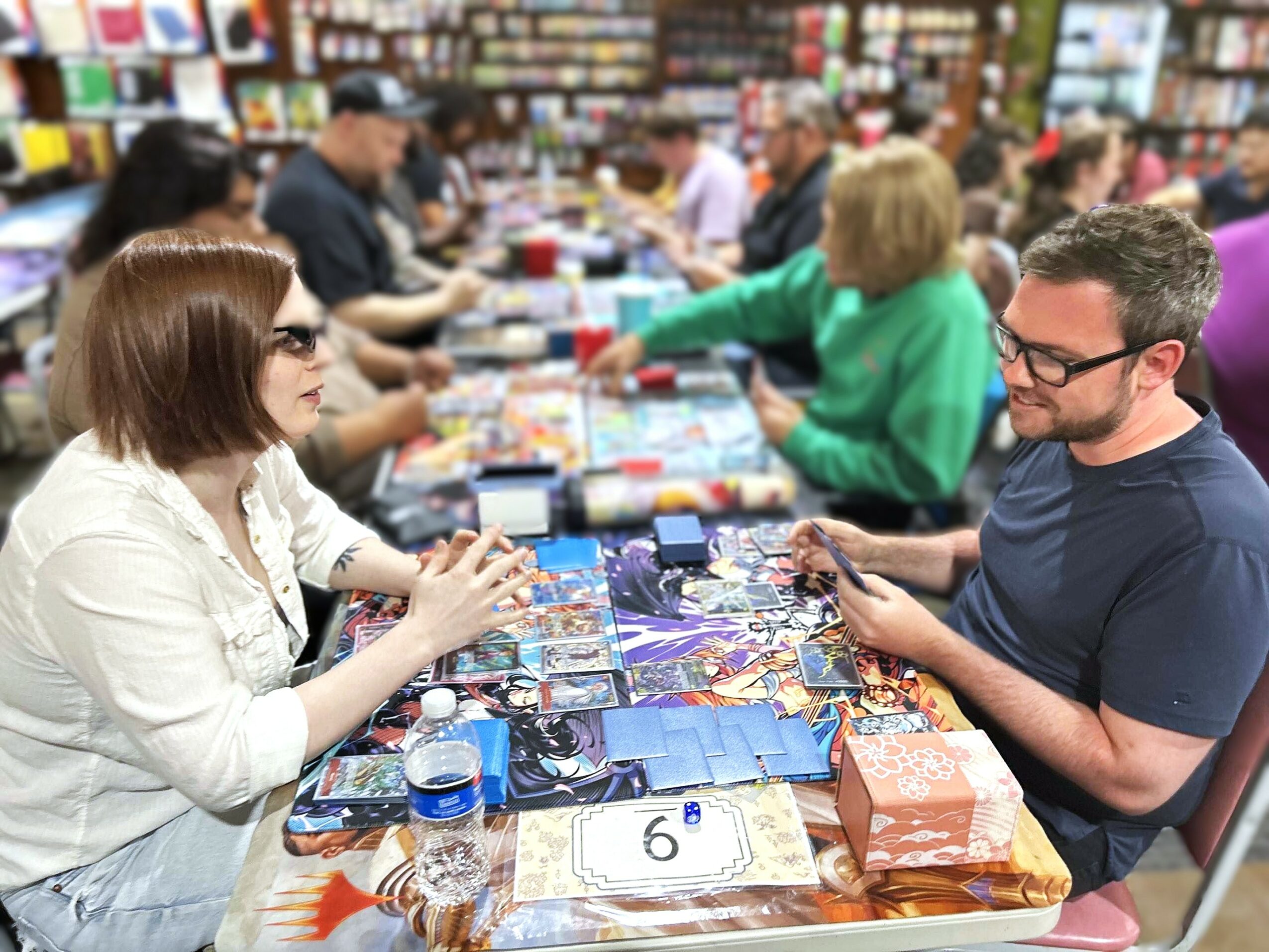 People playing board games at a table in a store, with shelves of products in the background.