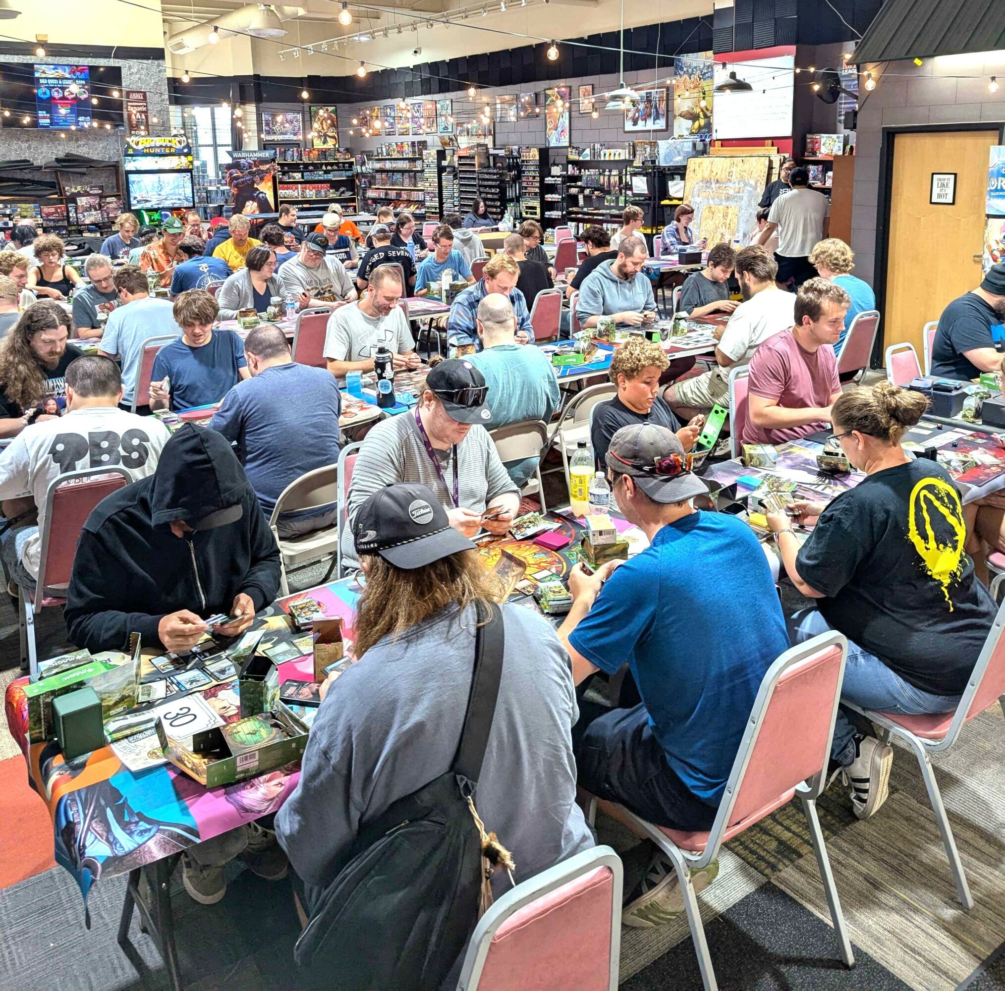People playing board games at tables in a crowded indoor space, with shelves and screens in the background.