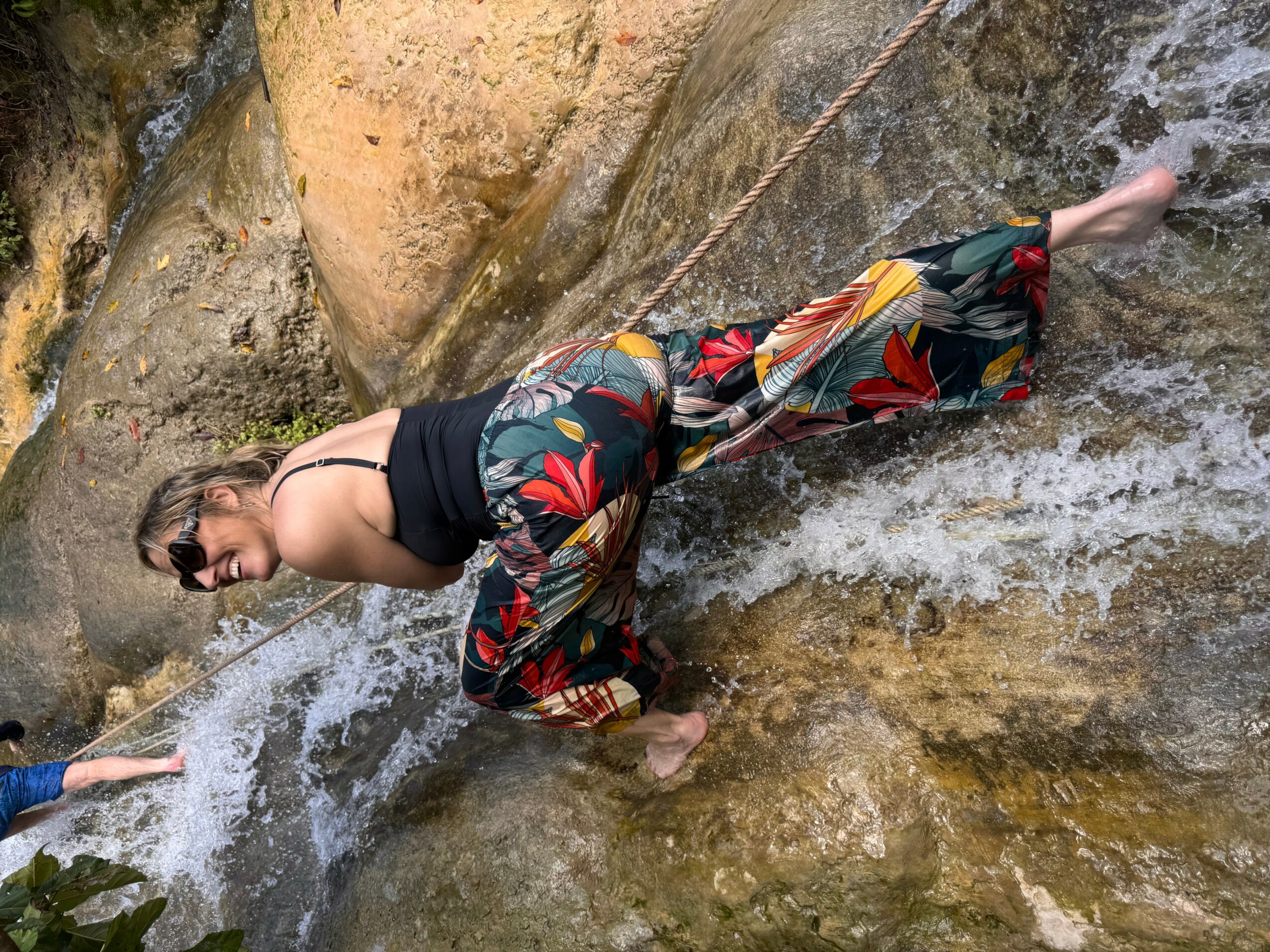 Person climbing wet rocks near water, wearing colorful pants and a black top, smiling, with a rocky background.
