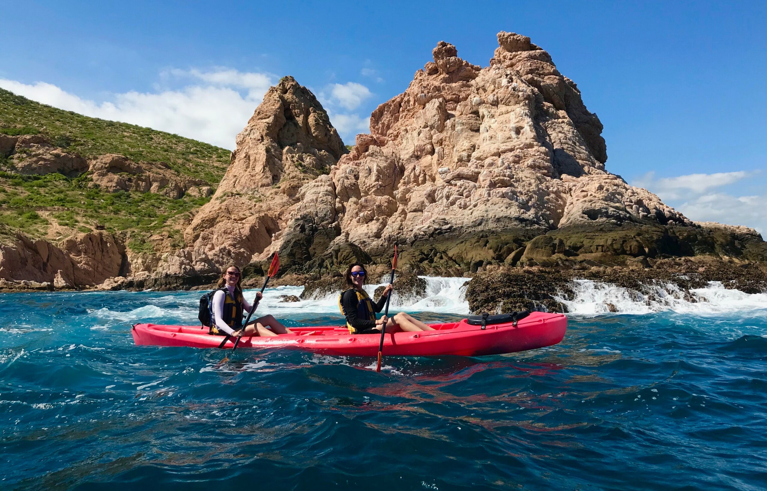 Two people kayaking near rocky cliffs with blue water and sky, wearing life jackets and holding paddles.