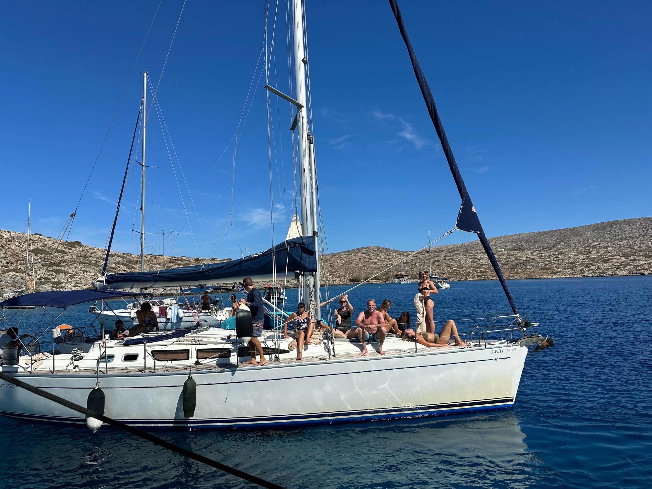 People relaxing on a sailboat with a blue sky and water, and hills in the background.