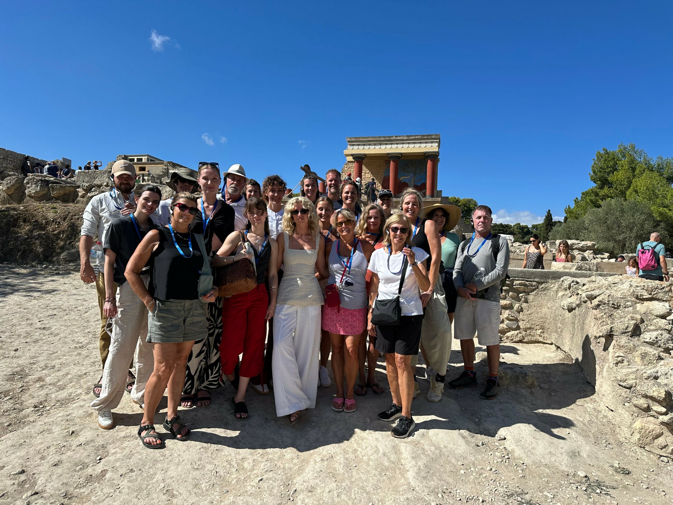 Group of people standing outdoors on a sunny day with ancient ruins and a building in the background.