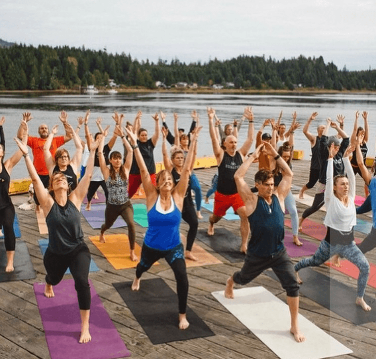 Group of people practicing yoga outdoors on a wooden deck near water, with trees in the background, raising arms in a pose.
