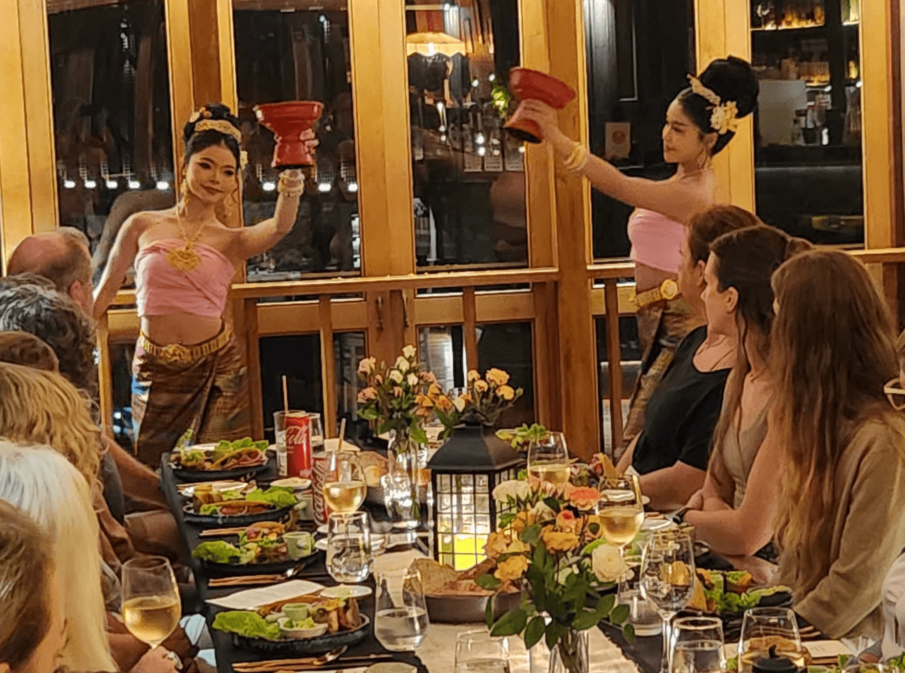 Two performers in traditional costumes dance with red hats in front of seated guests at a dinner event, with food and flowers on the table.