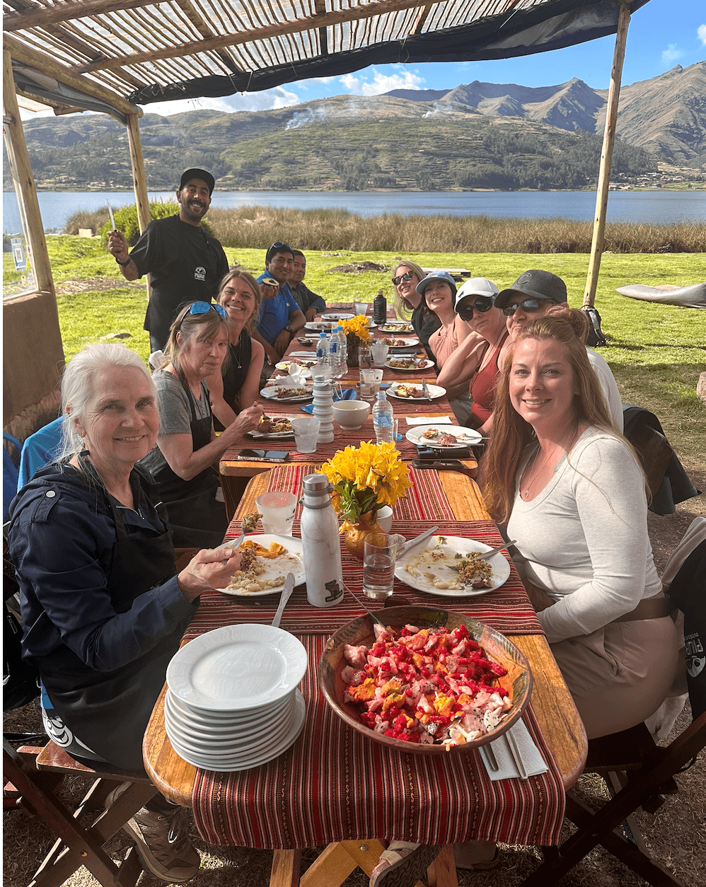 Group of people dining outdoors at a long table with mountains and lake in background, under a shaded canopy, with food and flowers.