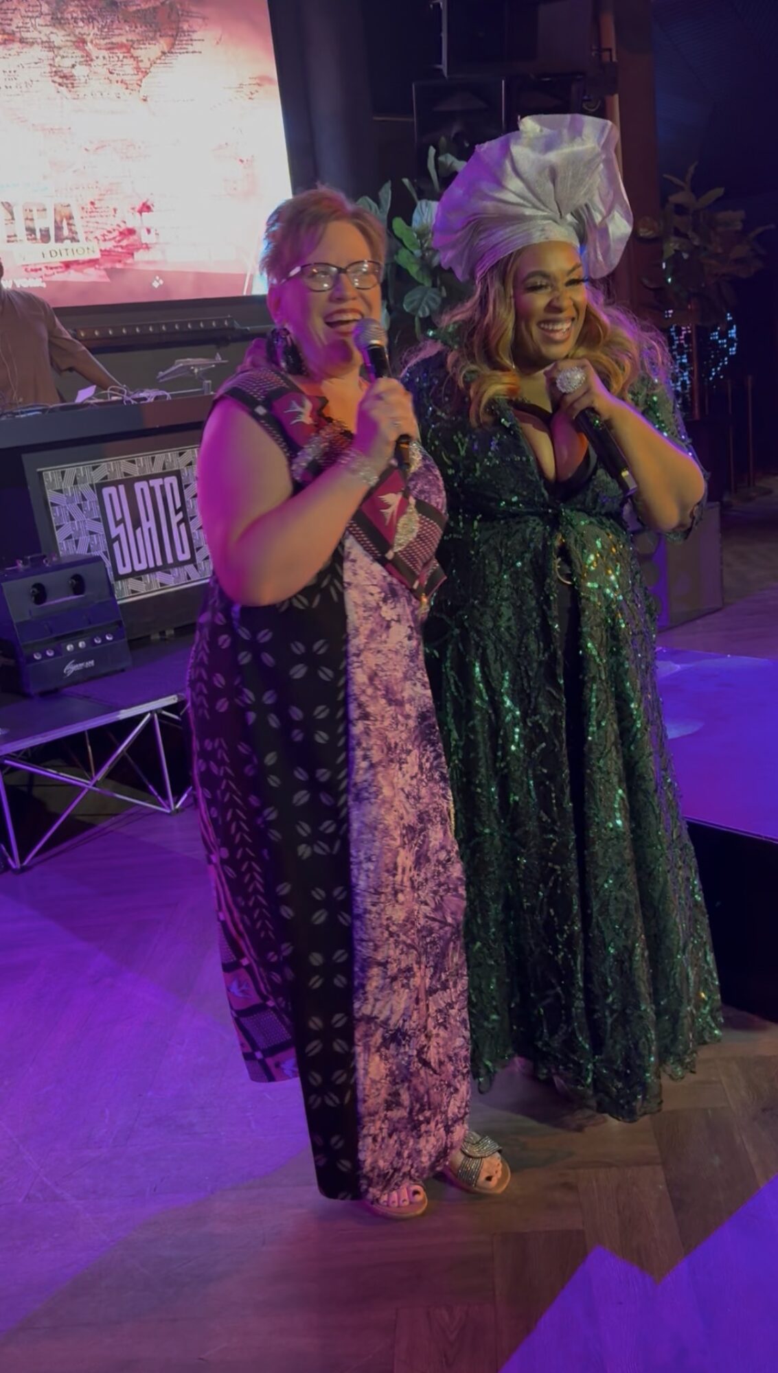 Two women in colorful dresses holding microphones on stage, smiling, with a screen and musical equipment behind them.