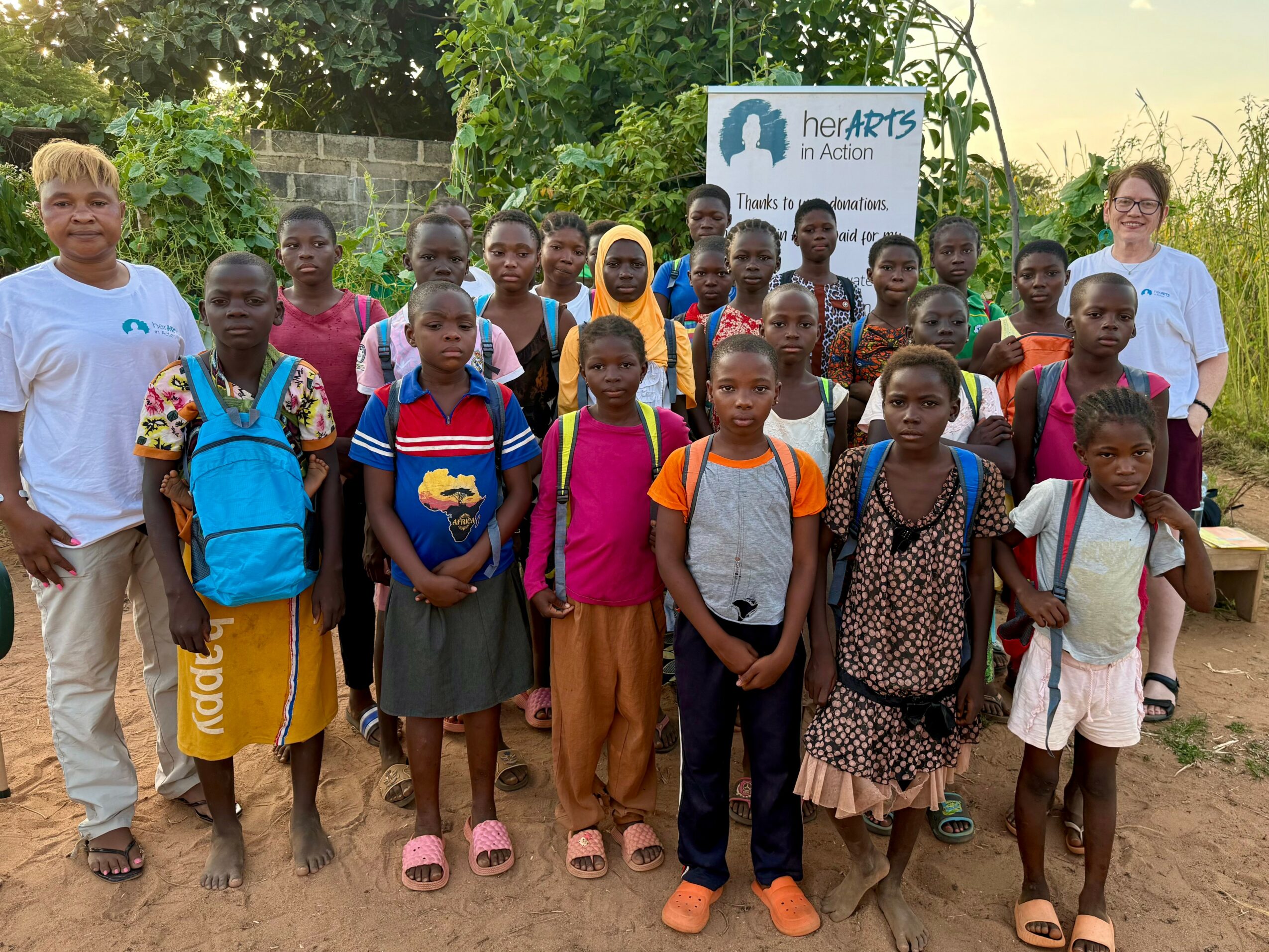 Group of children and two adults outdoors, some with backpacks, in front of a sign for HemArts in Action.