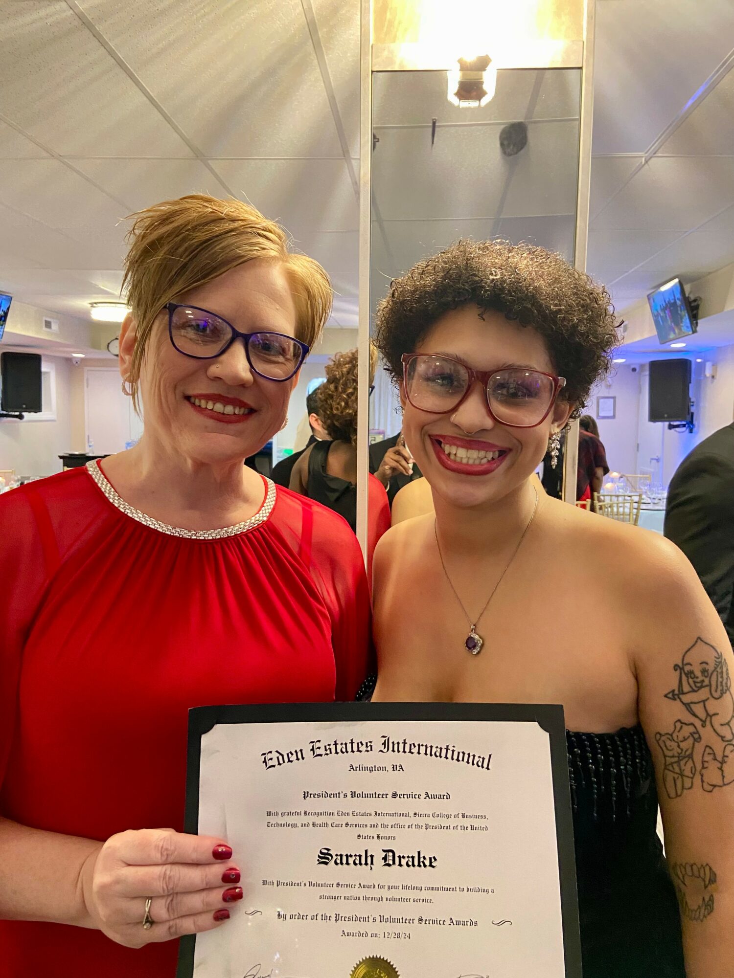 Two women smiling, one holding a certificate, in an indoor setting with bright lighting and a mirror behind them.