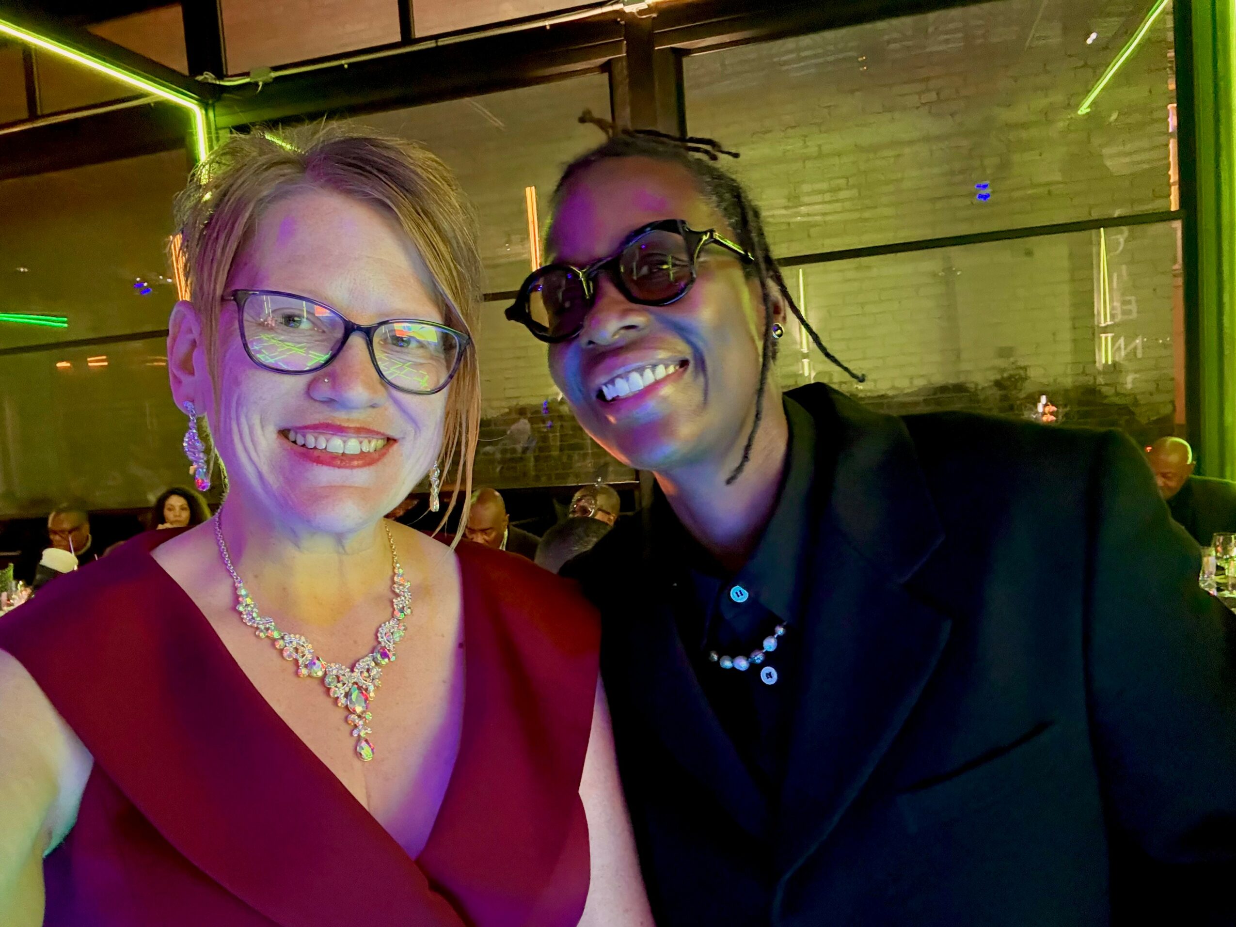 Two women smiling at a social event, one wearing glasses and a red dress, the other with sunglasses and a black outfit.