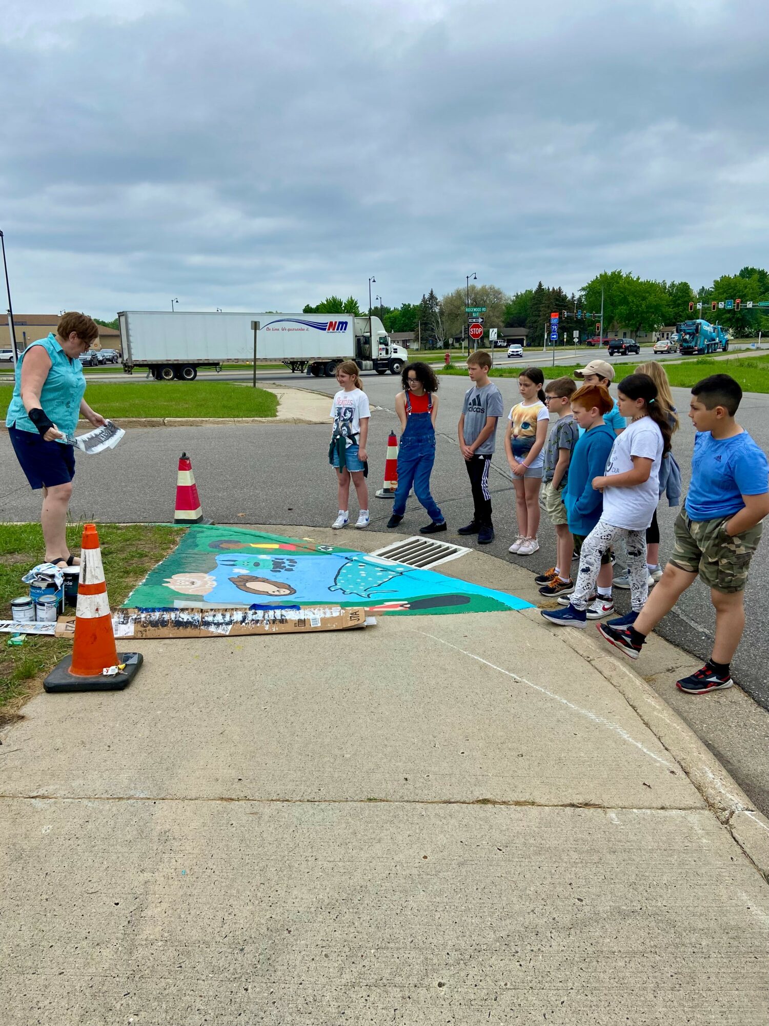 Group of children watching a woman paint a large colorful mural on the ground outdoors, with traffic cones nearby.