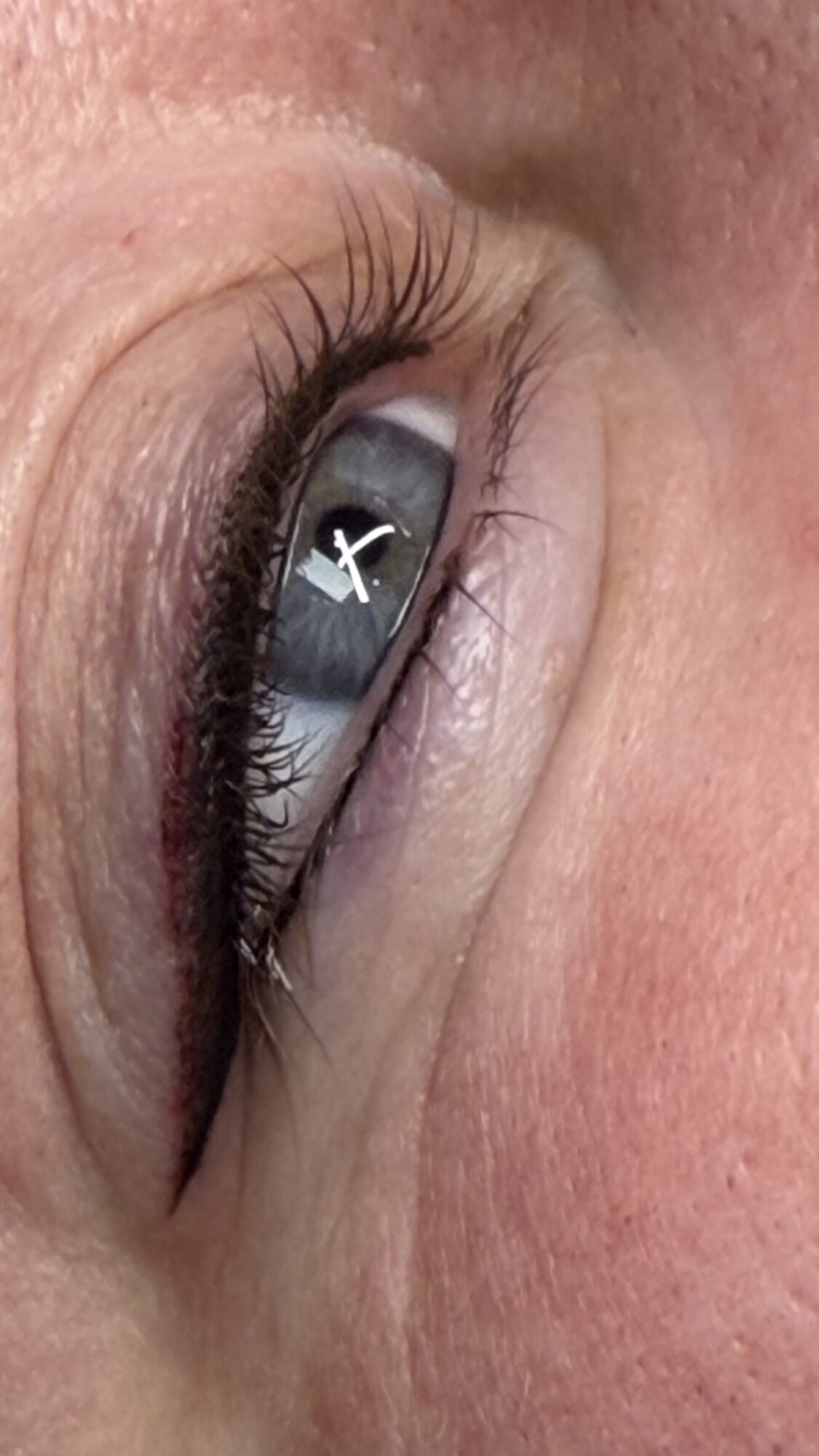 Close-up of a person's eye with visible eyelashes and reflection in the cornea, showing a white cross and a dark background.