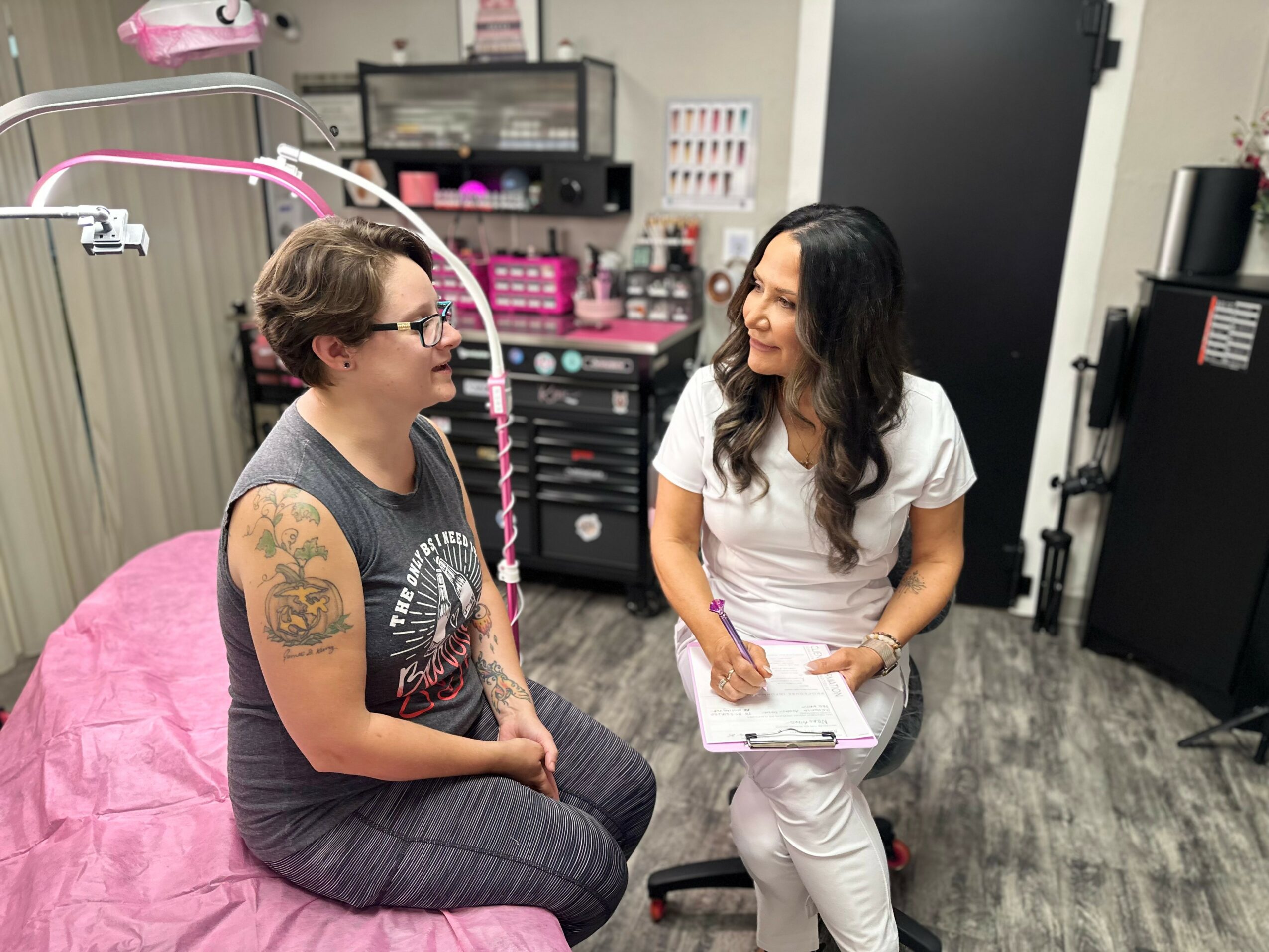 Two women sit and talk in a medical or beauty clinic, one on a pink bed and the other on a stool, with equipment in the background.
