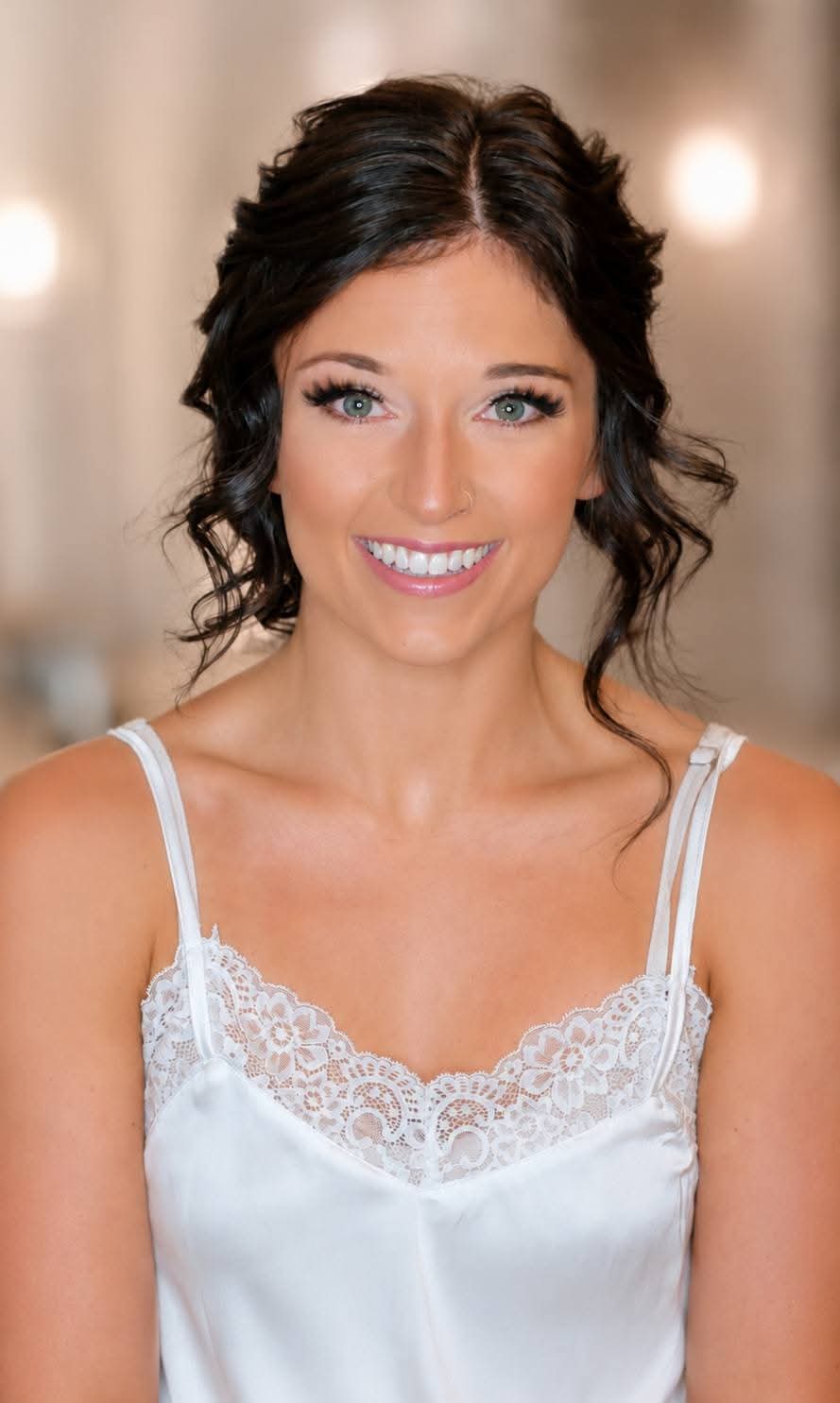 Smiling woman with dark wavy hair in a white lace-trimmed top, indoors with warm lighting