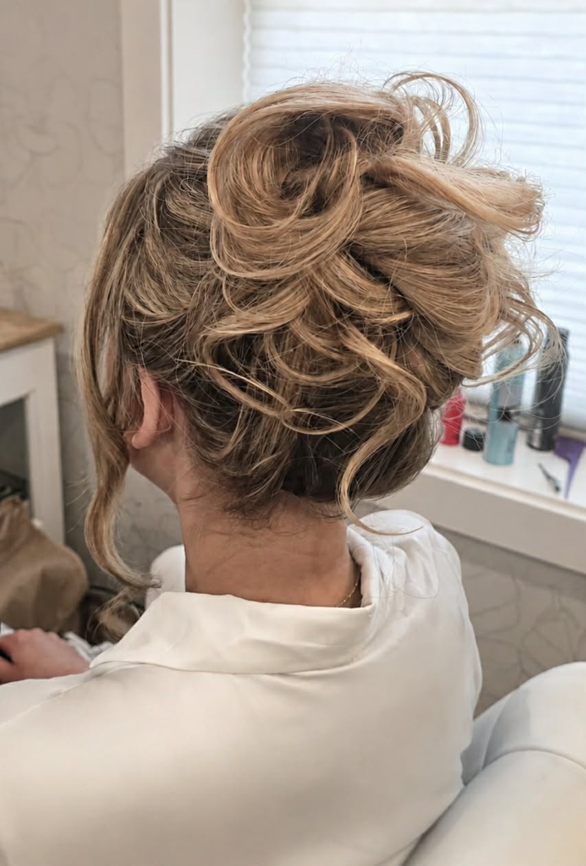 Back view of a woman with styled, voluminous hair in a salon, wearing a white cape, near a window with beauty products.