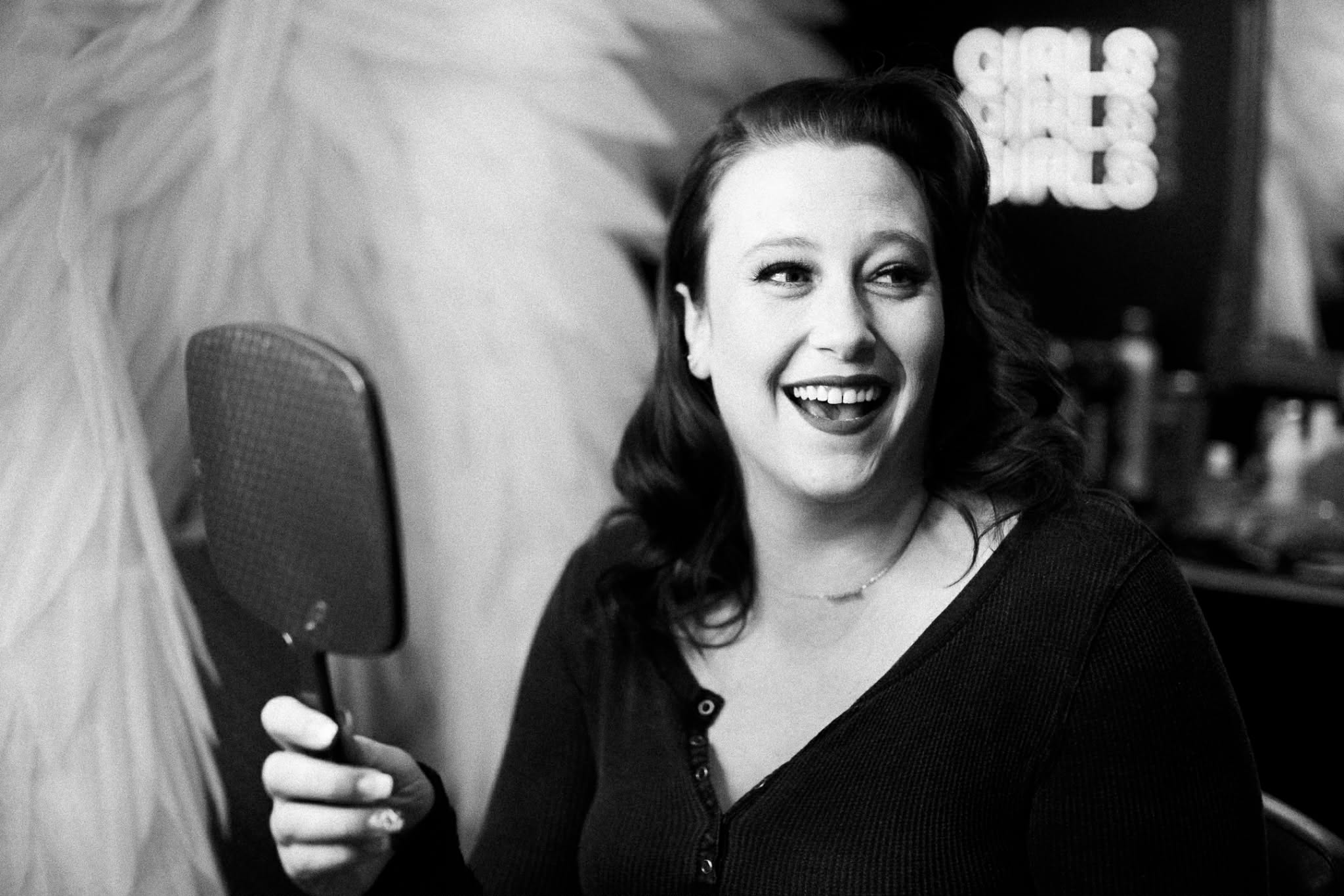 Woman with dark hair smiling and holding a mirror, in a room with textured wall and blurred background.