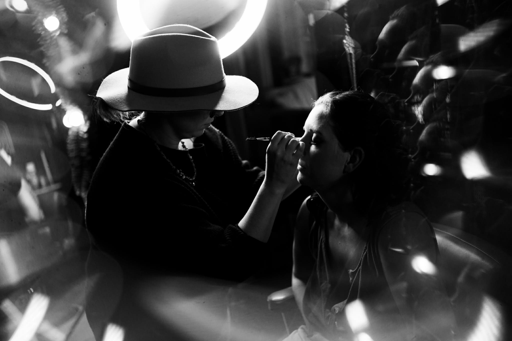Two women in a dimly lit setting, one applying makeup or face paint to the other, with blurred lights in the background.