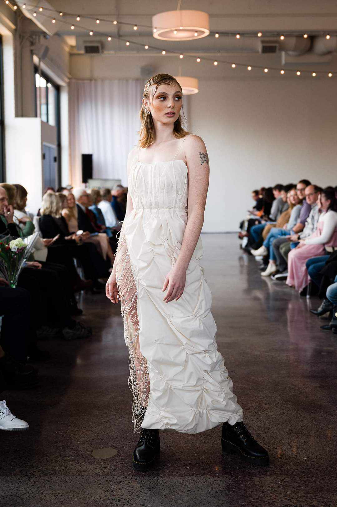 Female model walking on runway in white dress with lace details, audience seated on both sides, indoor event setting.