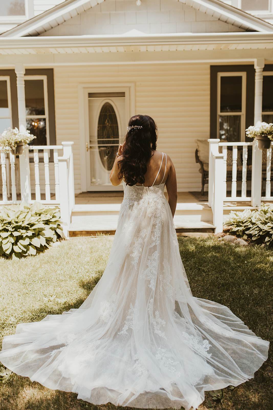 Woman in wedding dress standing on grass in front of a house with porch, facing away from the camera.